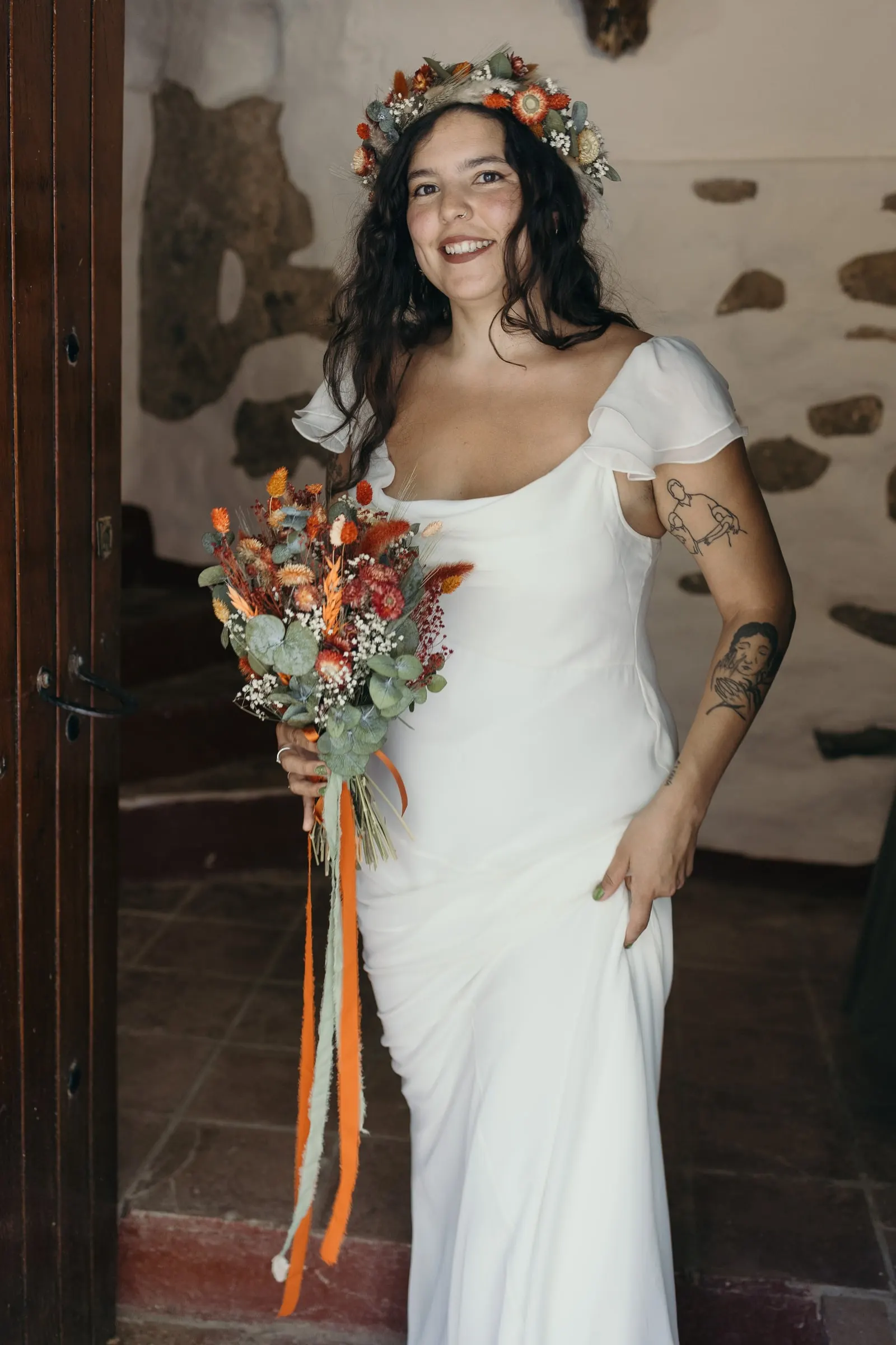 Bride in a doorway with a stone wall, holding a bouquet with orange ribbons