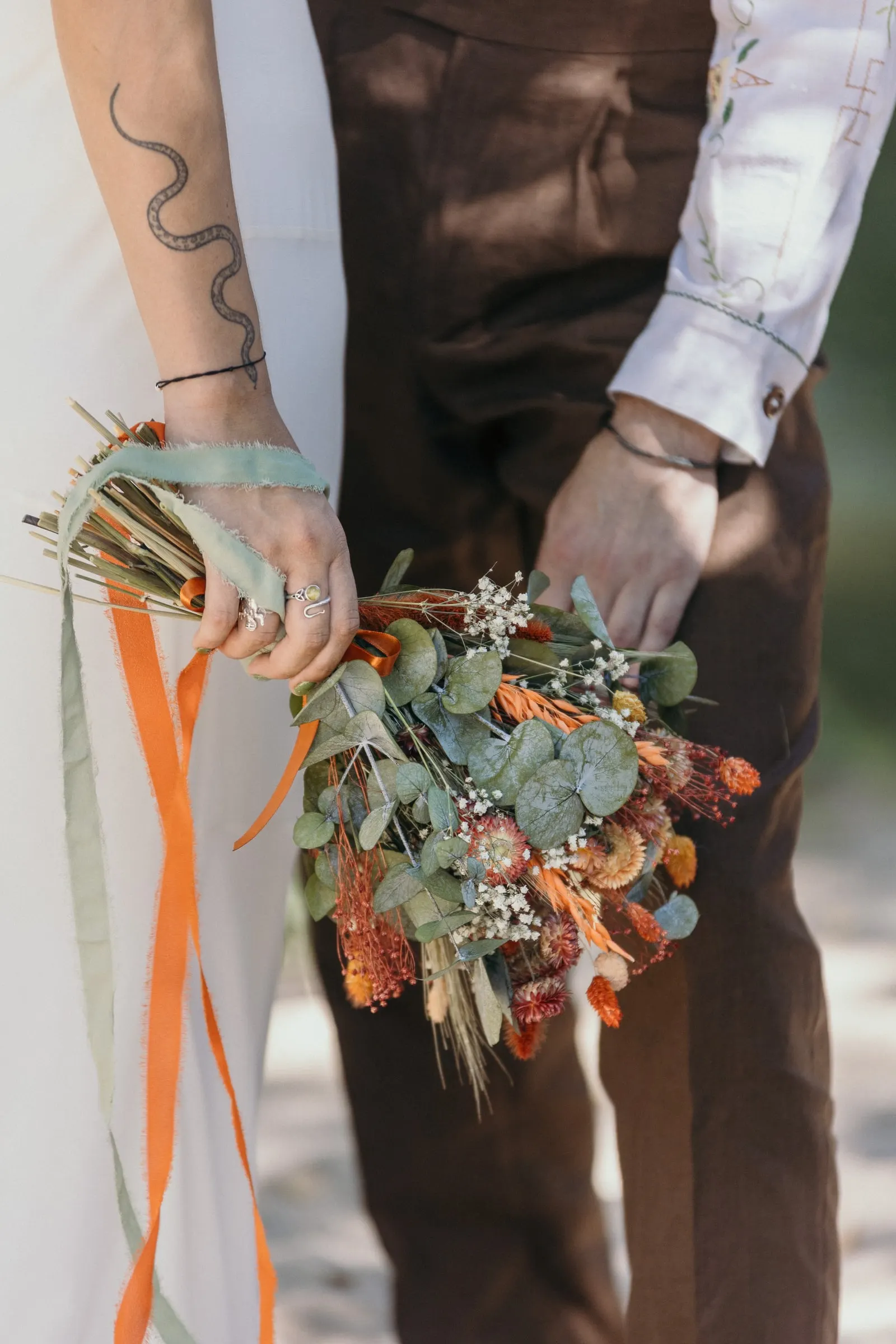 Bride's tattooed hand holding a dried-flower bouquet with long ribbons