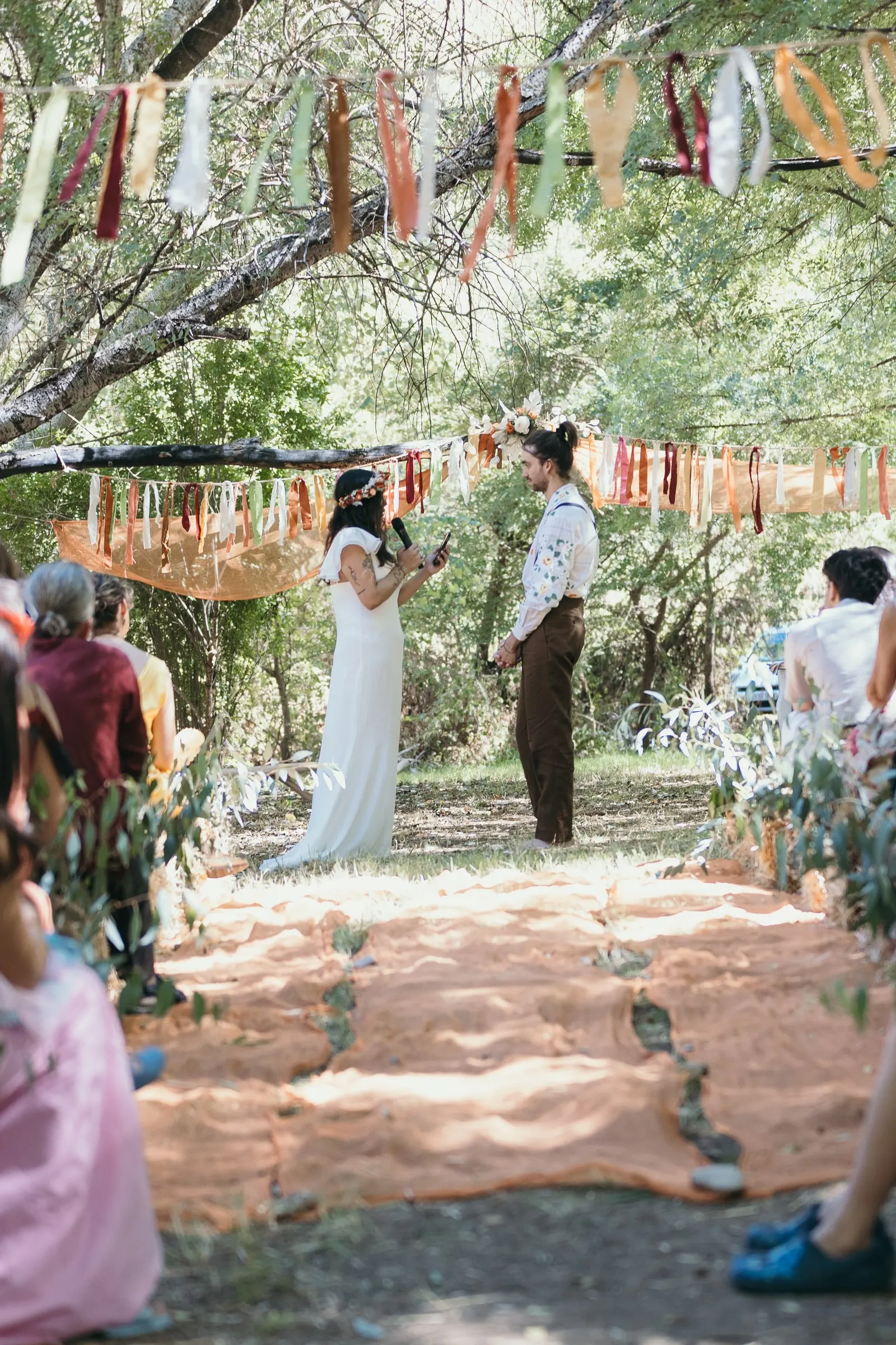 Wide view of the ceremony at Molino La Flor: terracotta rug aisle and ribbon garlands in the trees