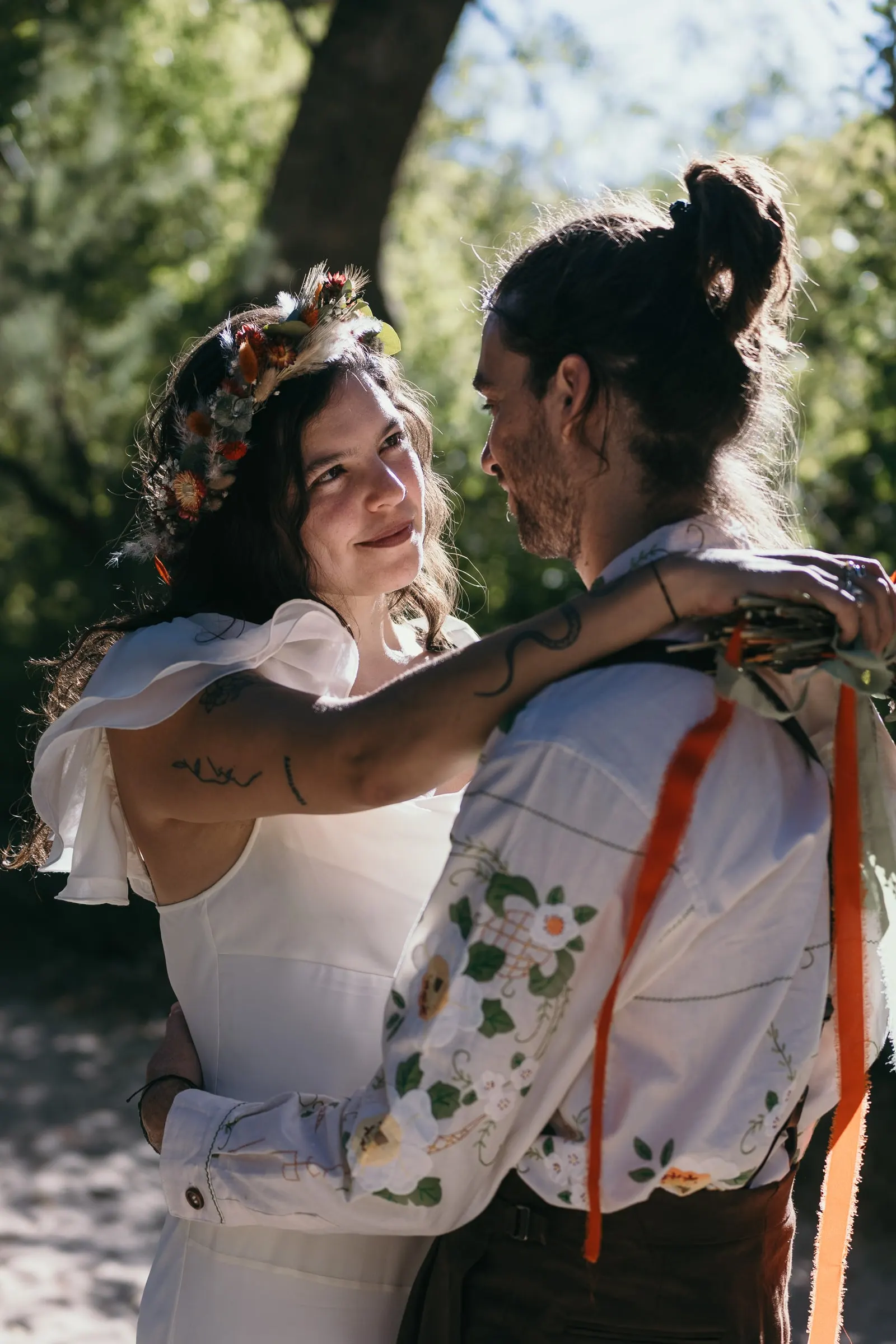 Bride with her arm around the groom's neck in the shade of the trees