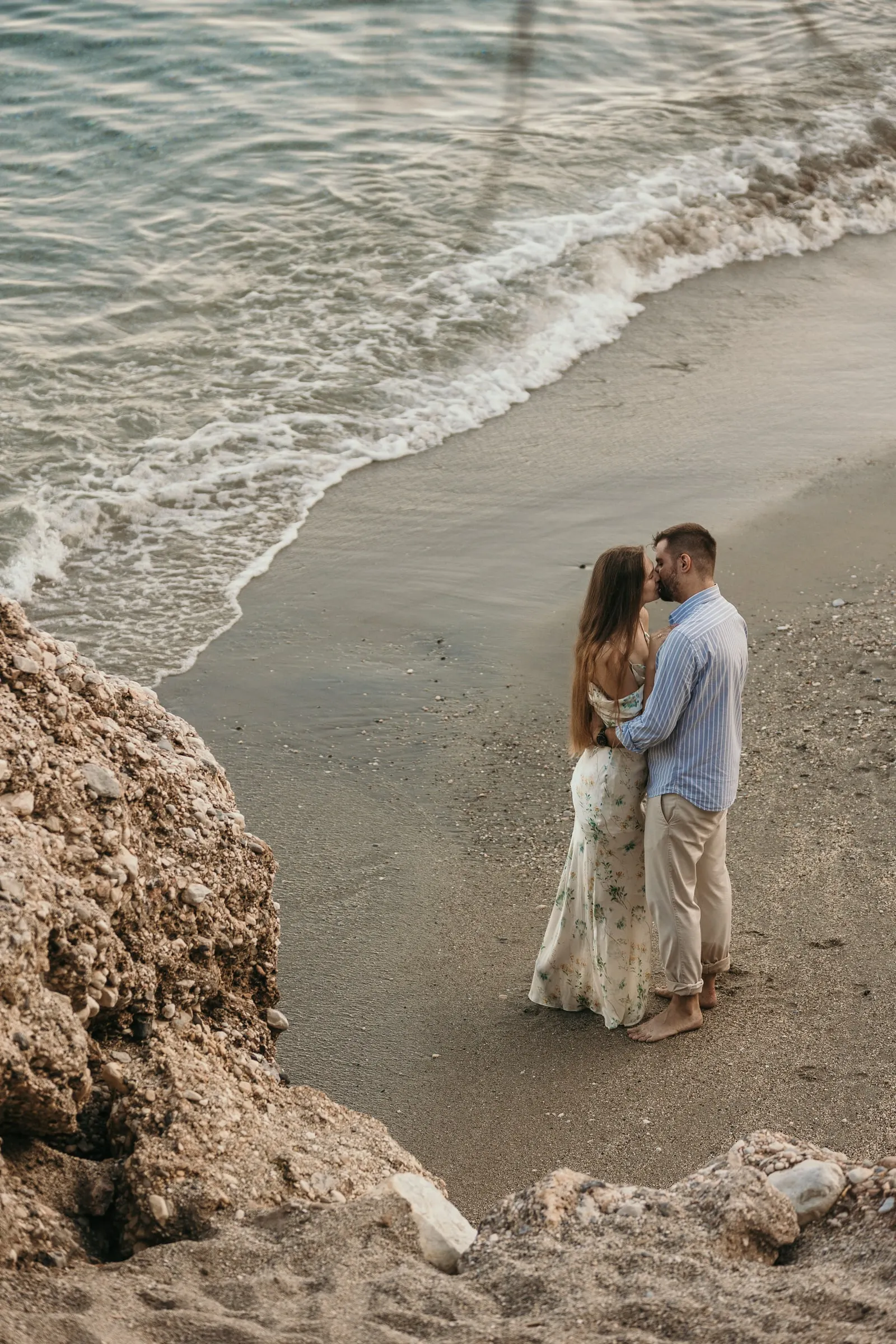 Couple embracing at the water's edge beside the rocks, seen from above, on the beach in Nerja