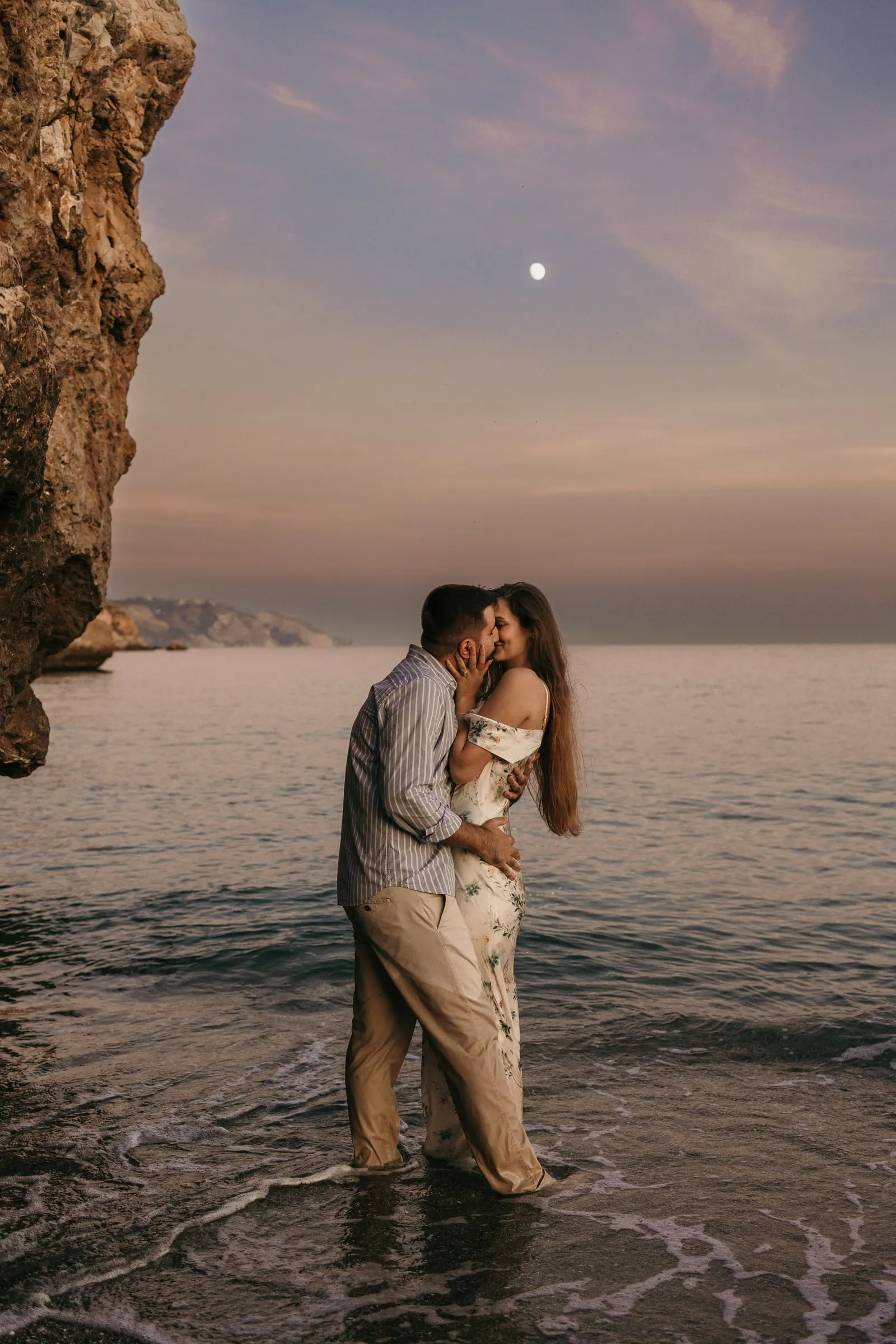 Couple kissing knee-deep in the sea at dusk, the moon over the cliff, on the beach in Nerja