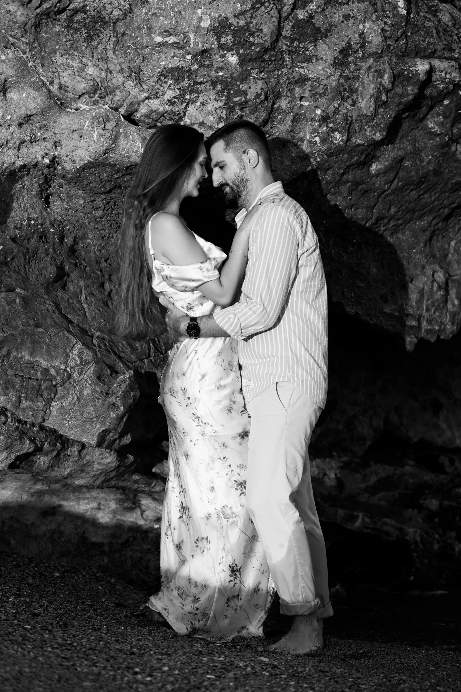 Black and white of the couple embracing in front of a dark rock on the beach in Nerja