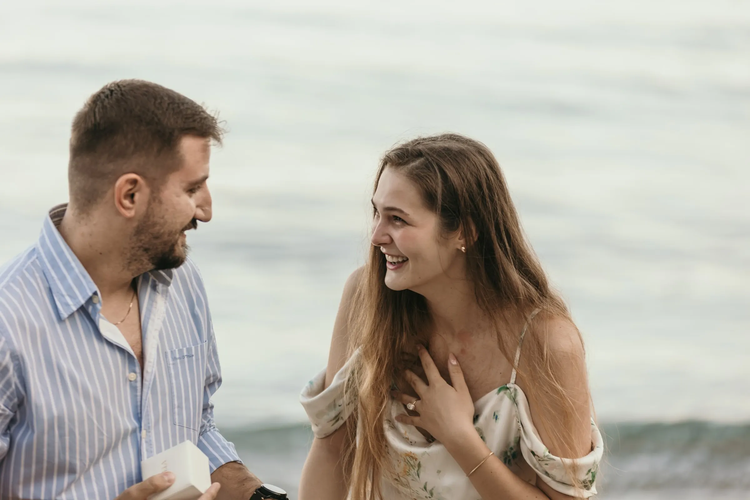 She laughs with her hand on her chest as he opens the ring box, on the beach in Nerja
