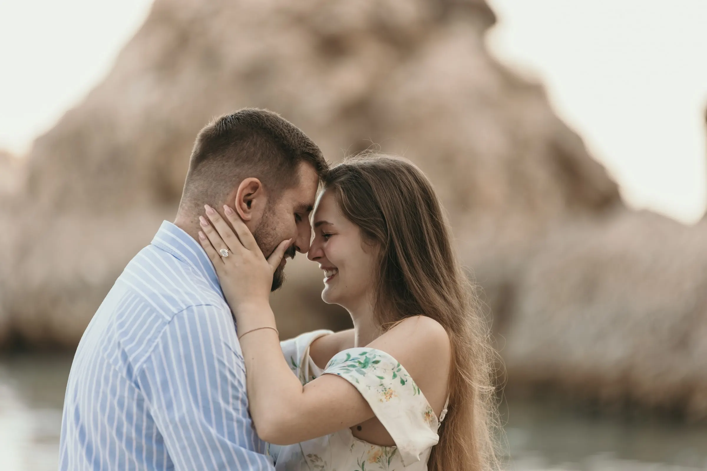 Couple touching foreheads, her ring visible, with the rocks behind, on the beach in Nerja