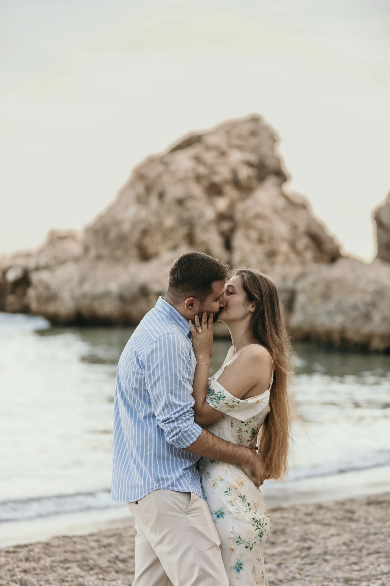 Couple kissing on the shingle with a large rock in the sea behind, on the beach in Nerja