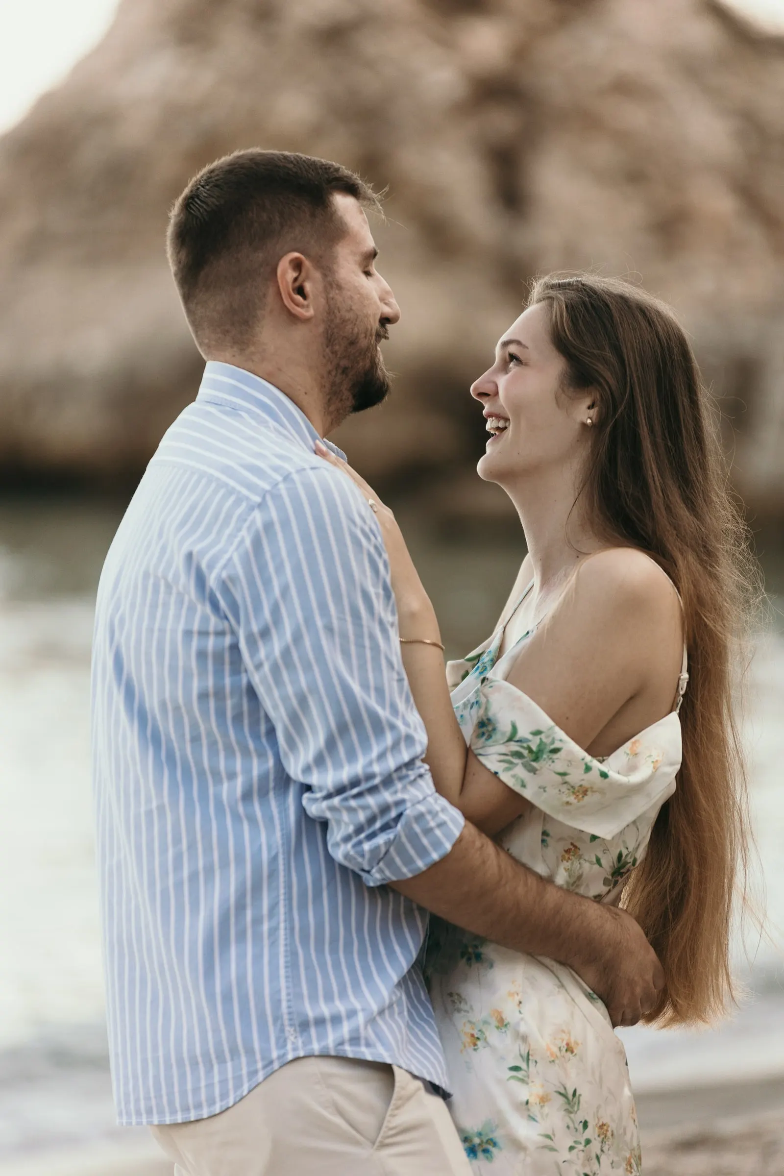 She looks up at him laughing as they embrace by the rocks, on the beach in Nerja