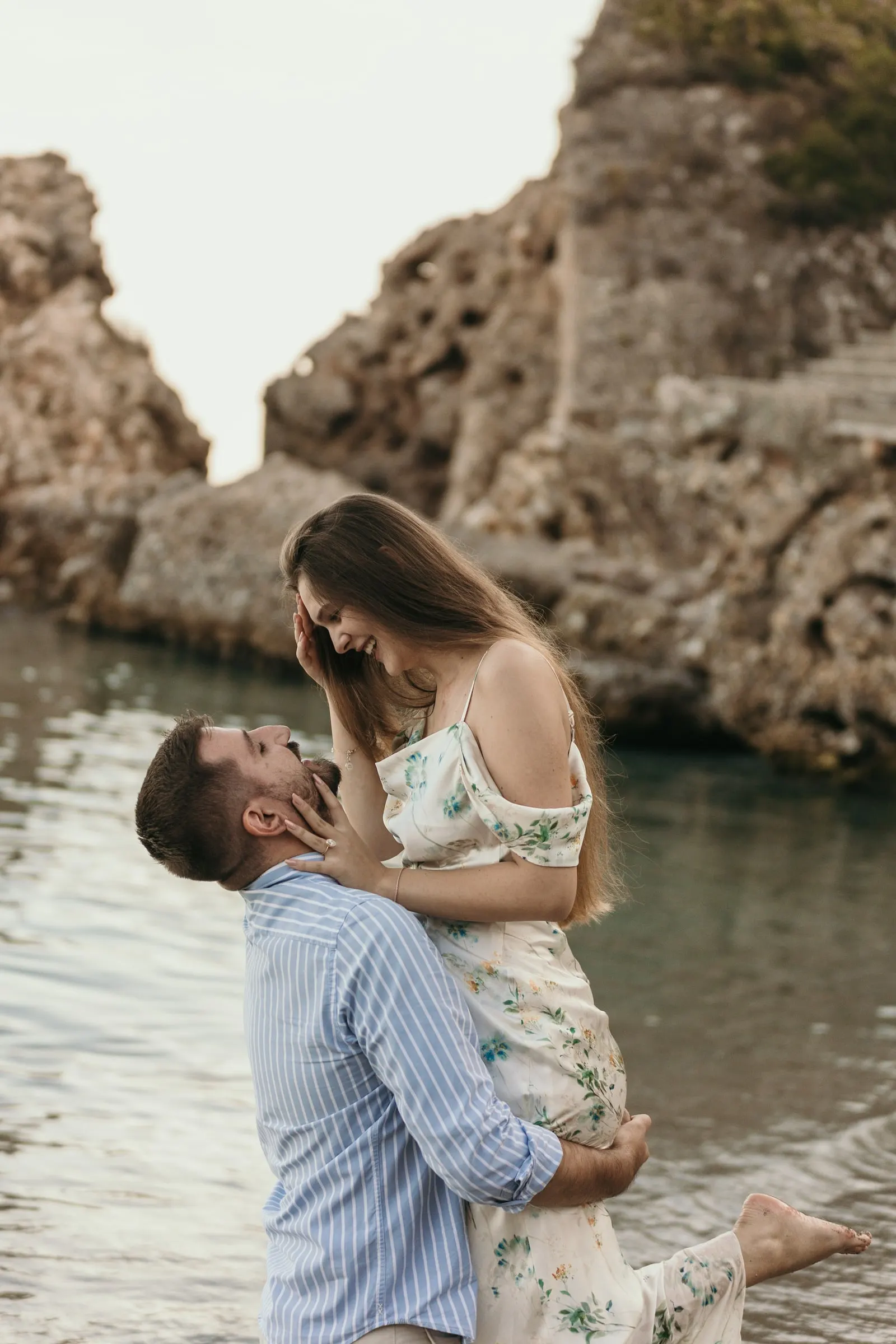 He lifts her up as she smiles down at him, with the rocky cove behind, on the beach in Nerja