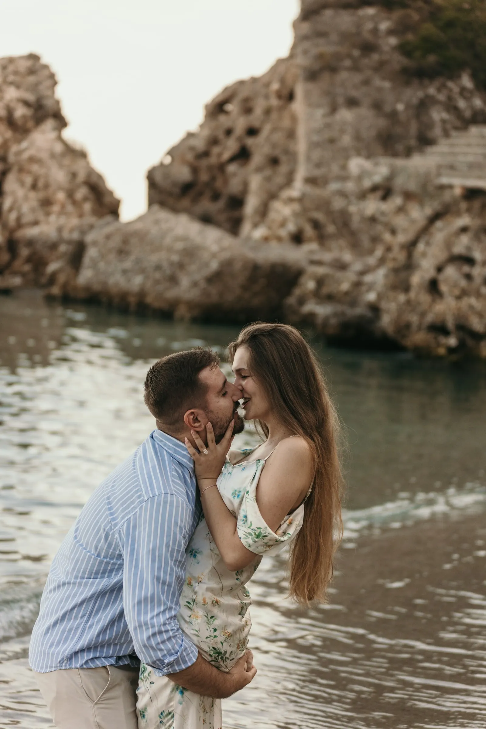 He lifts her for a kiss in the water in front of the rocky cove, on the beach in Nerja