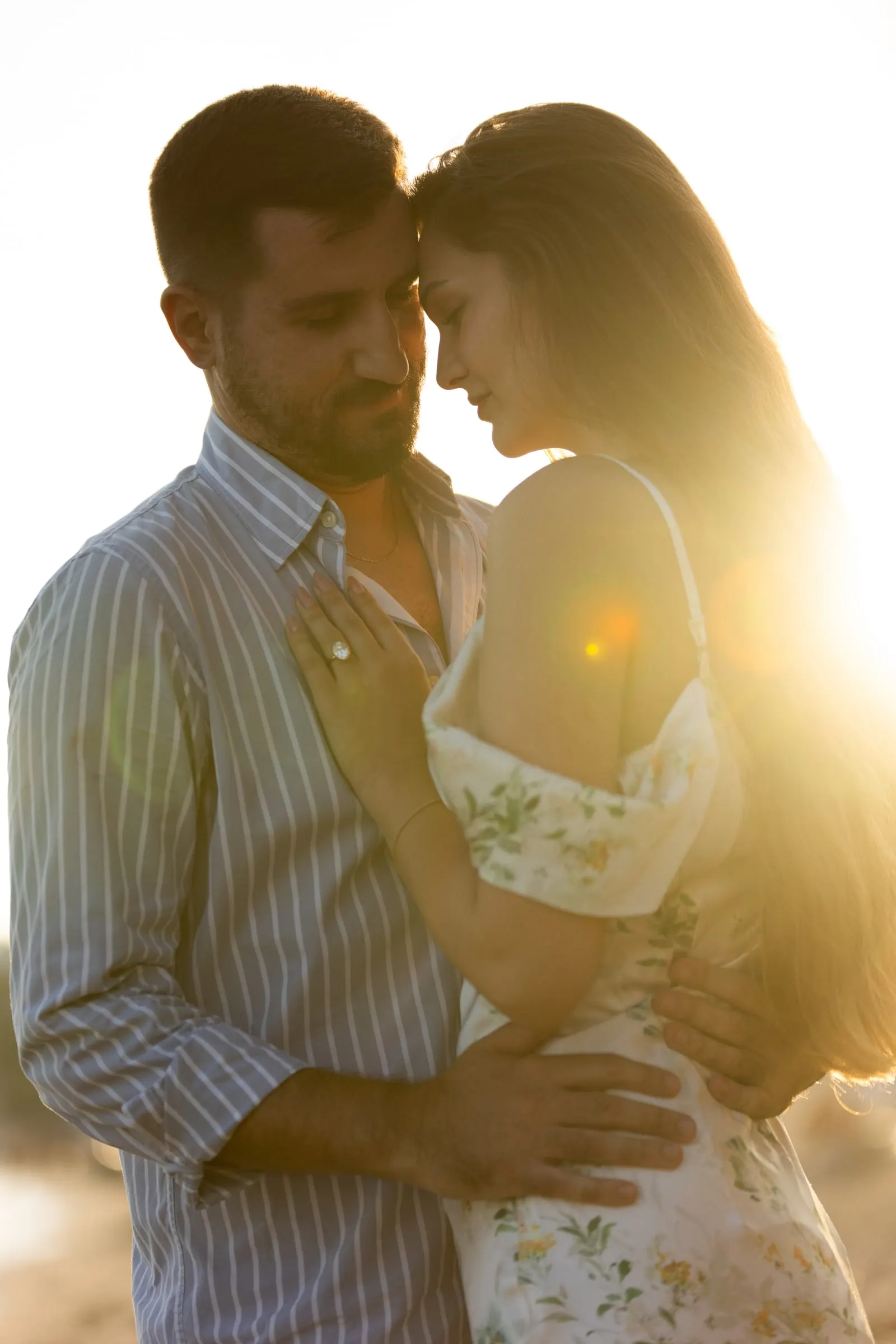 Couple forehead to forehead as the sun flares behind them, her ring visible, beach in Nerja