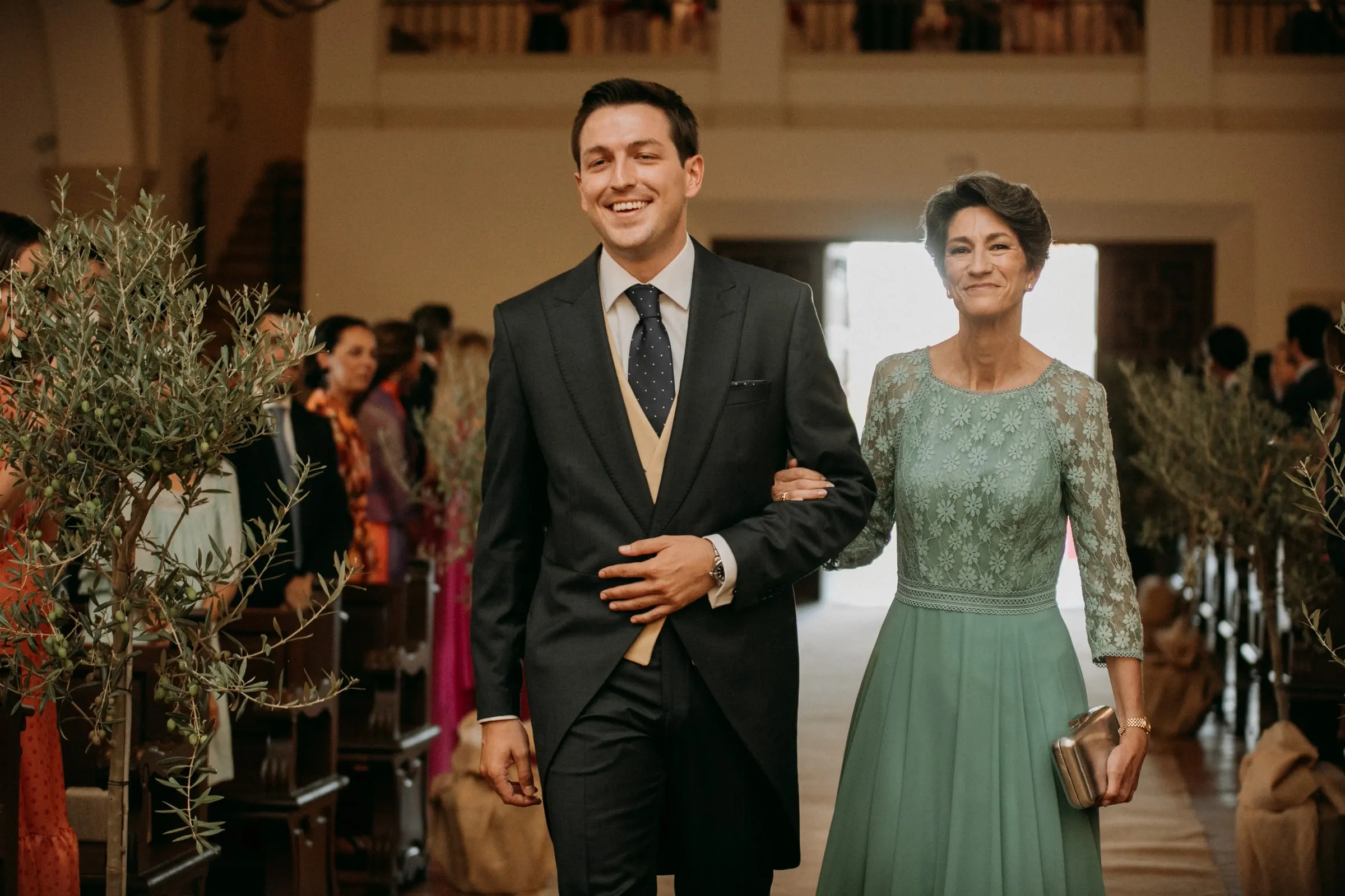 Groom and the woman in green walking up the aisle between guests and olive trees in the church