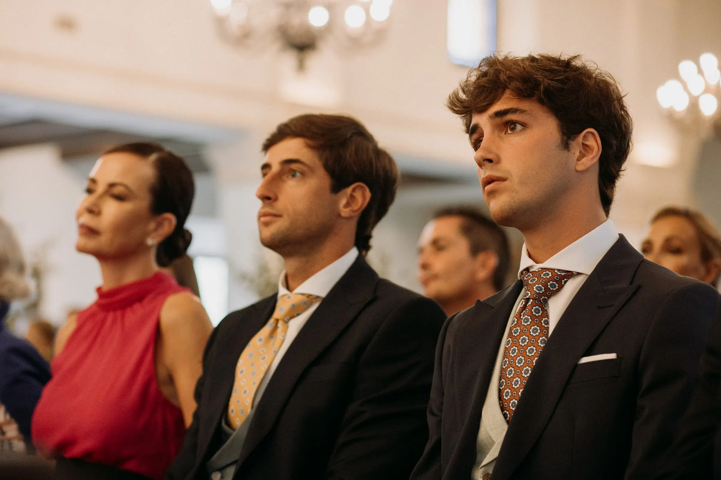 Young guests in suits and a woman in red watching the ceremony from the pews