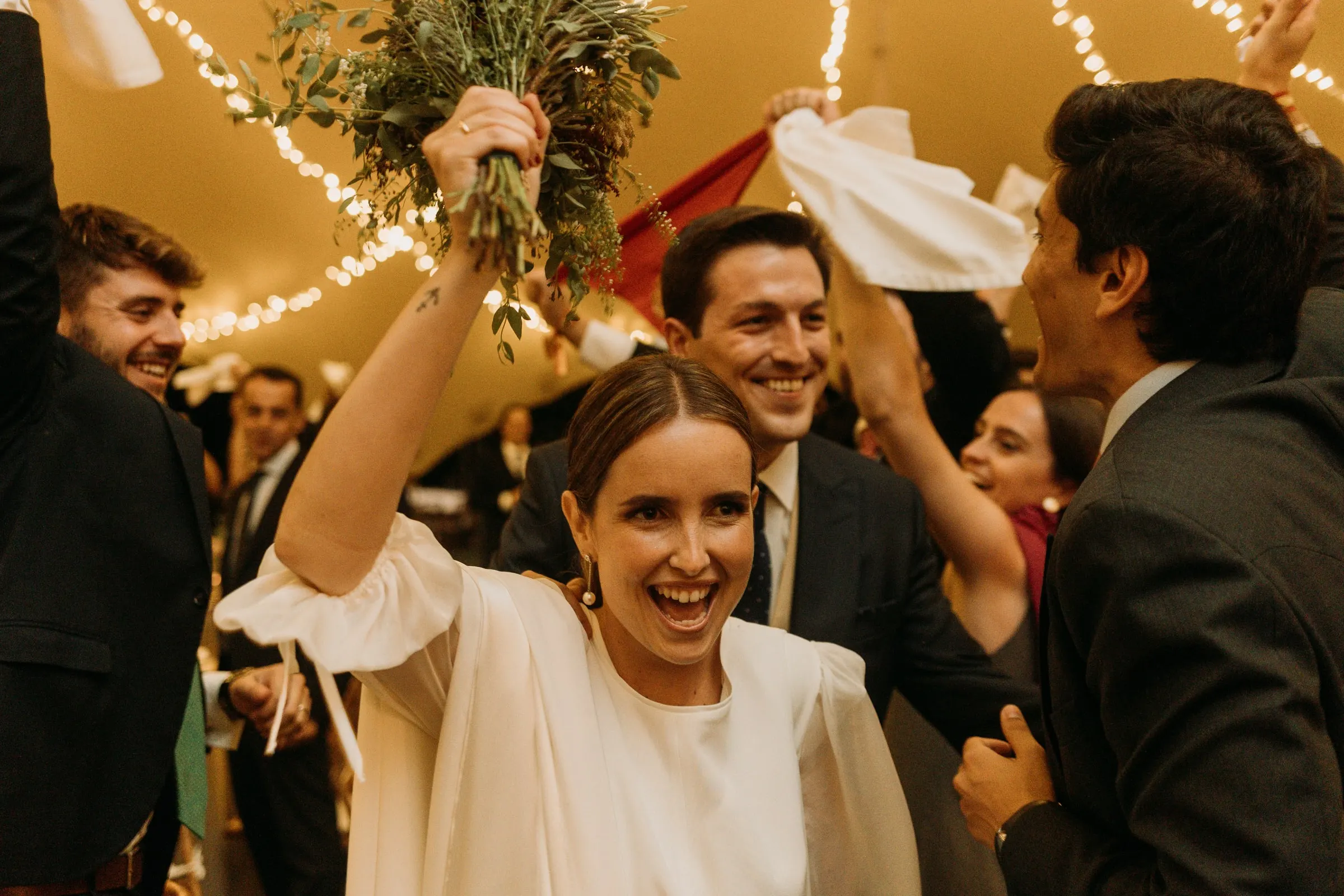Bride laughing as she lifts her bouquet among friends under the fairy lights