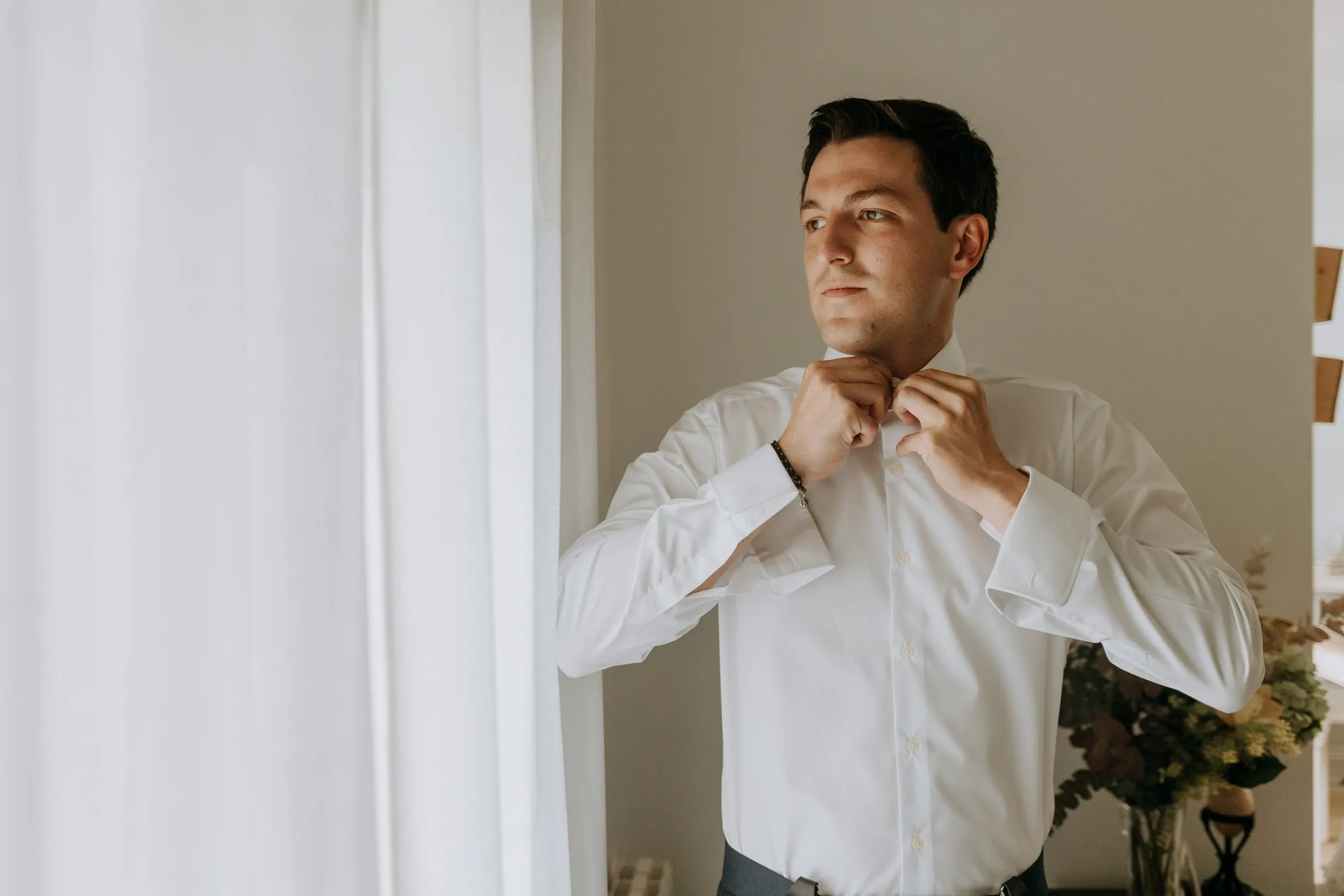Groom buttoning his white shirt by the window before the wedding in Sotogrande