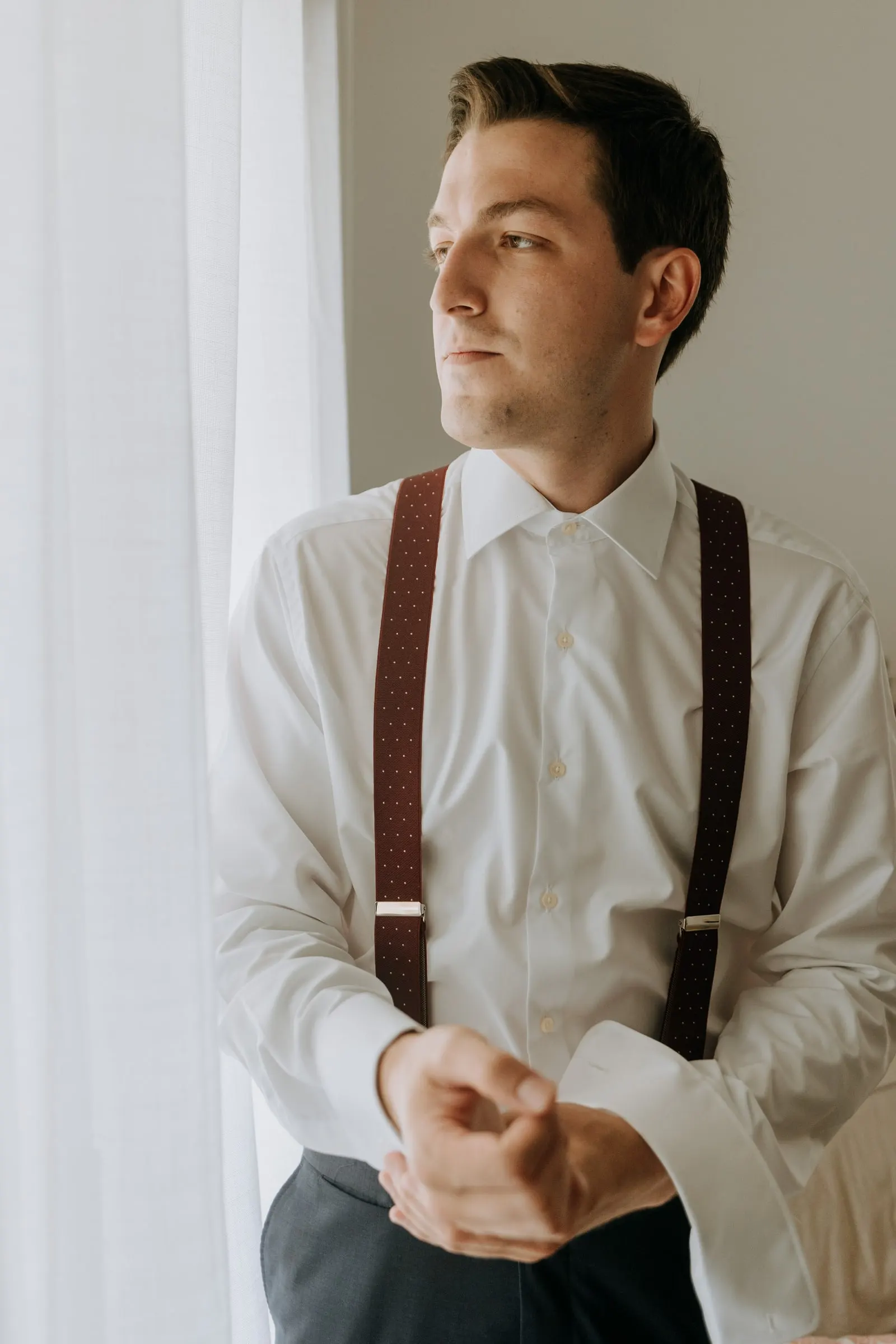 Groom in burgundy braces fastening his cuffs in the light of the window