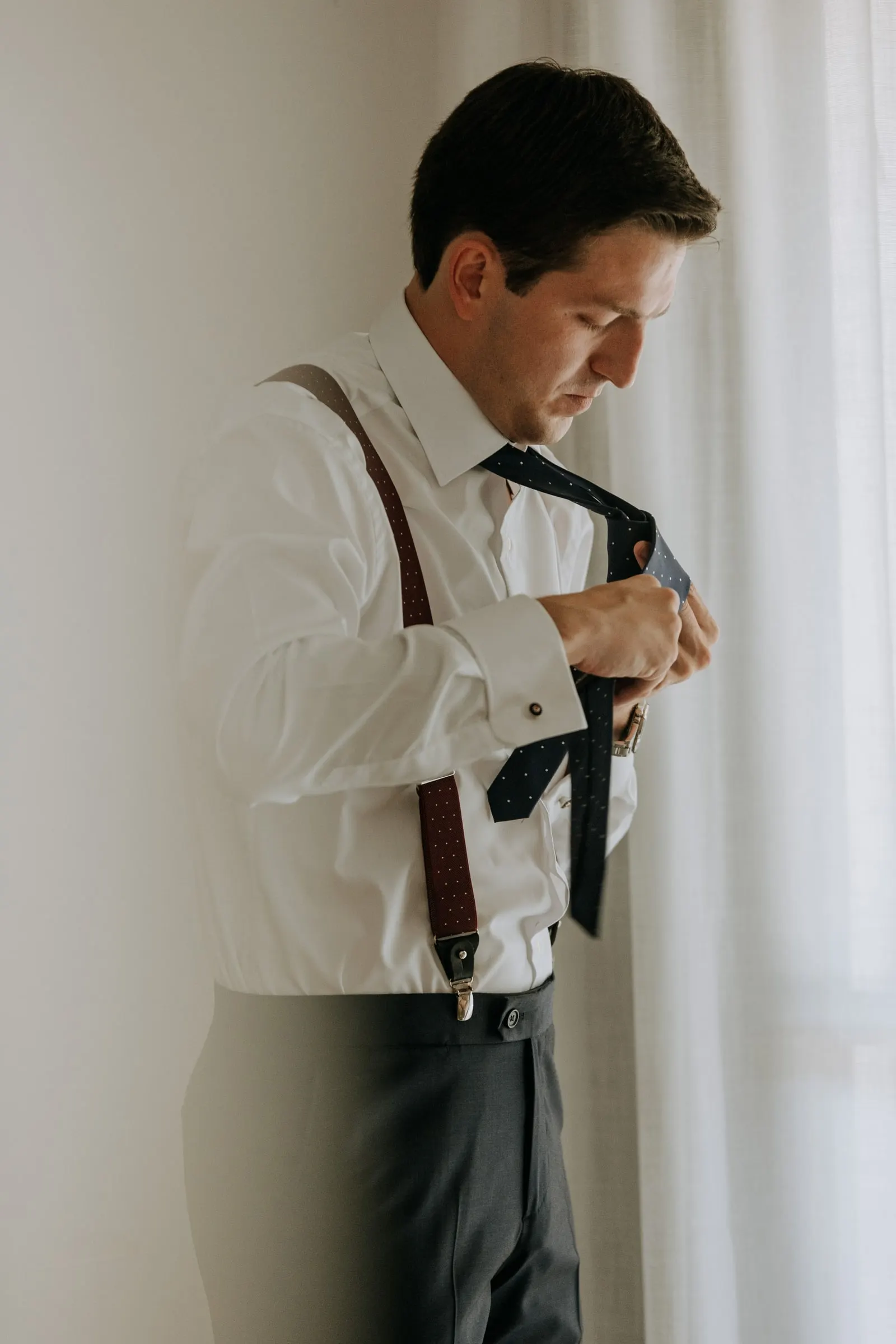 Groom tying his tie in front of the window
