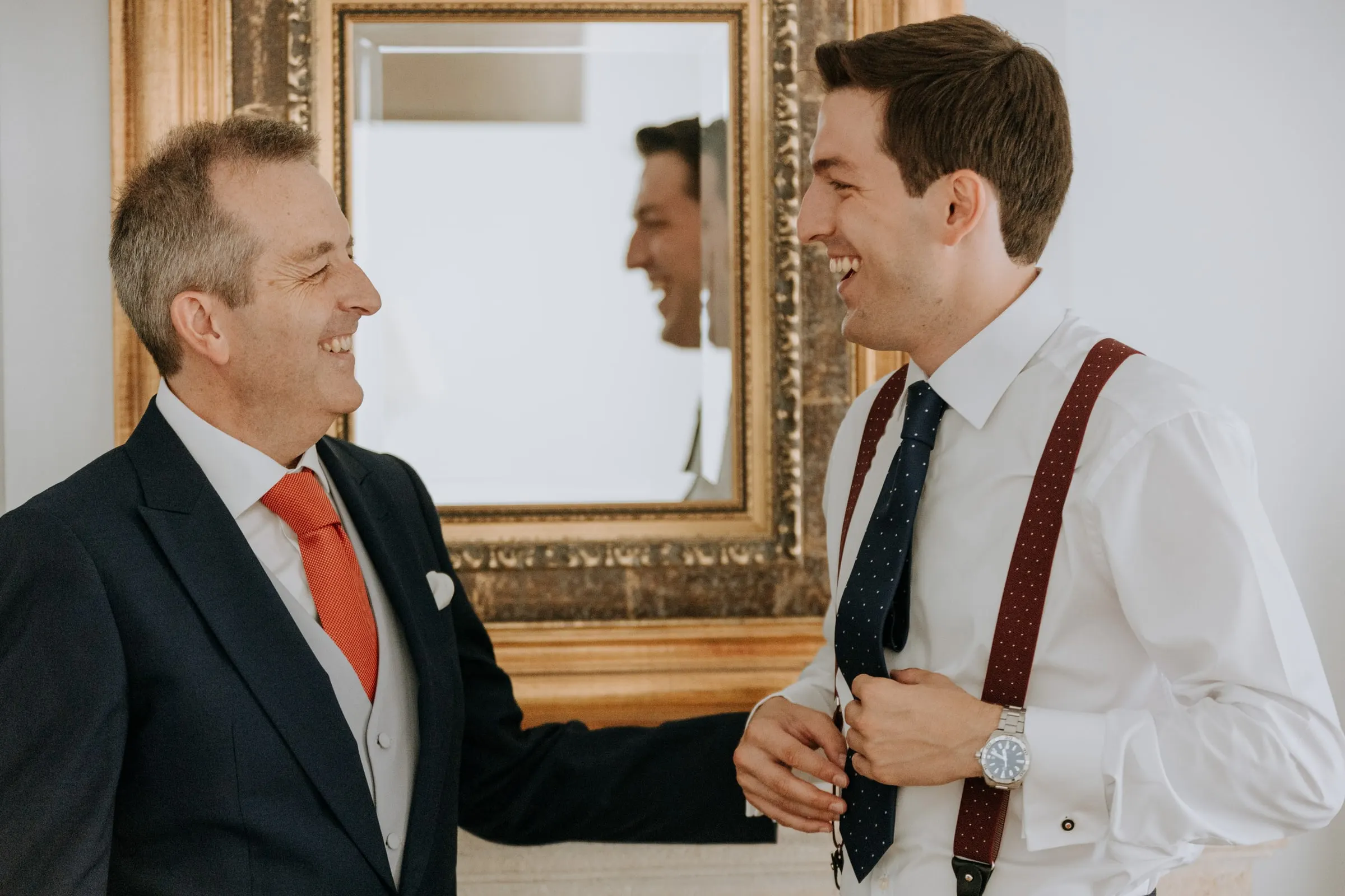 Groom sharing a laugh with an older guest in front of a gilded mirror