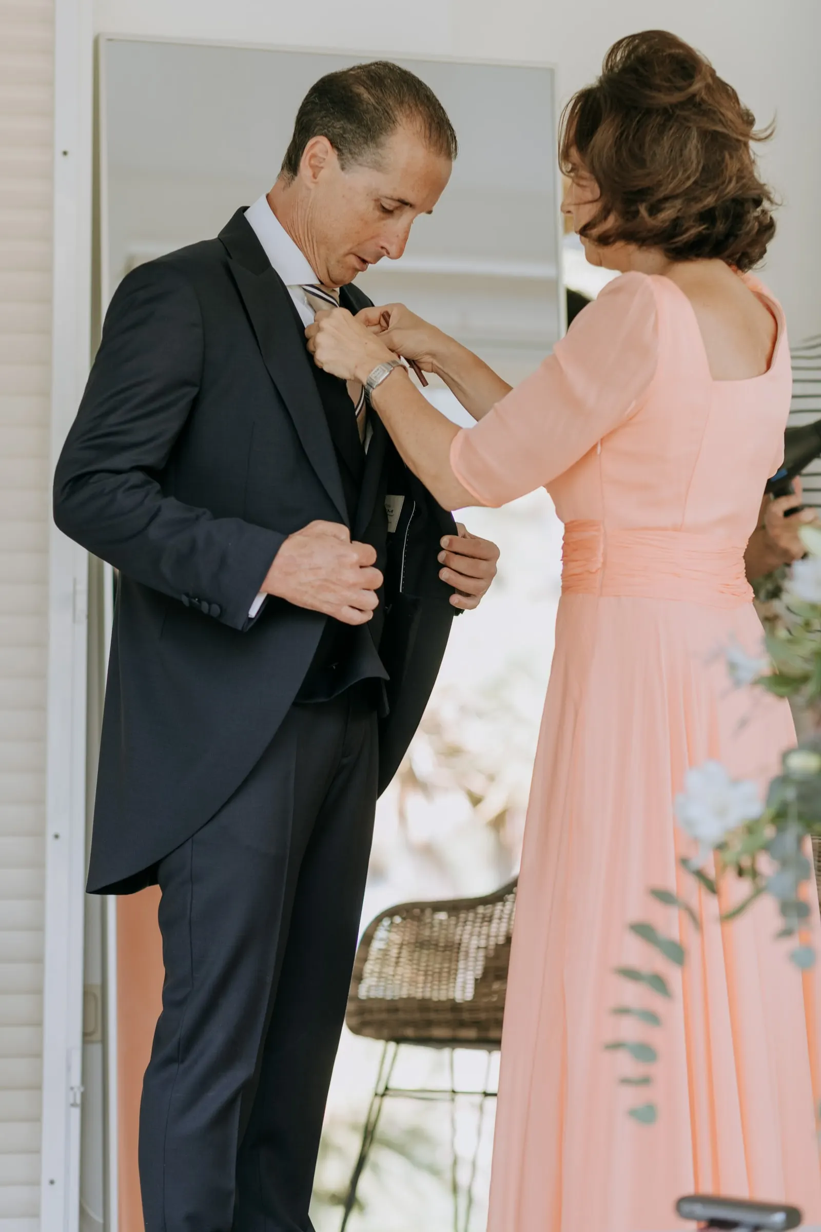 Woman in peach pinning the boutonniere on the groom's morning coat