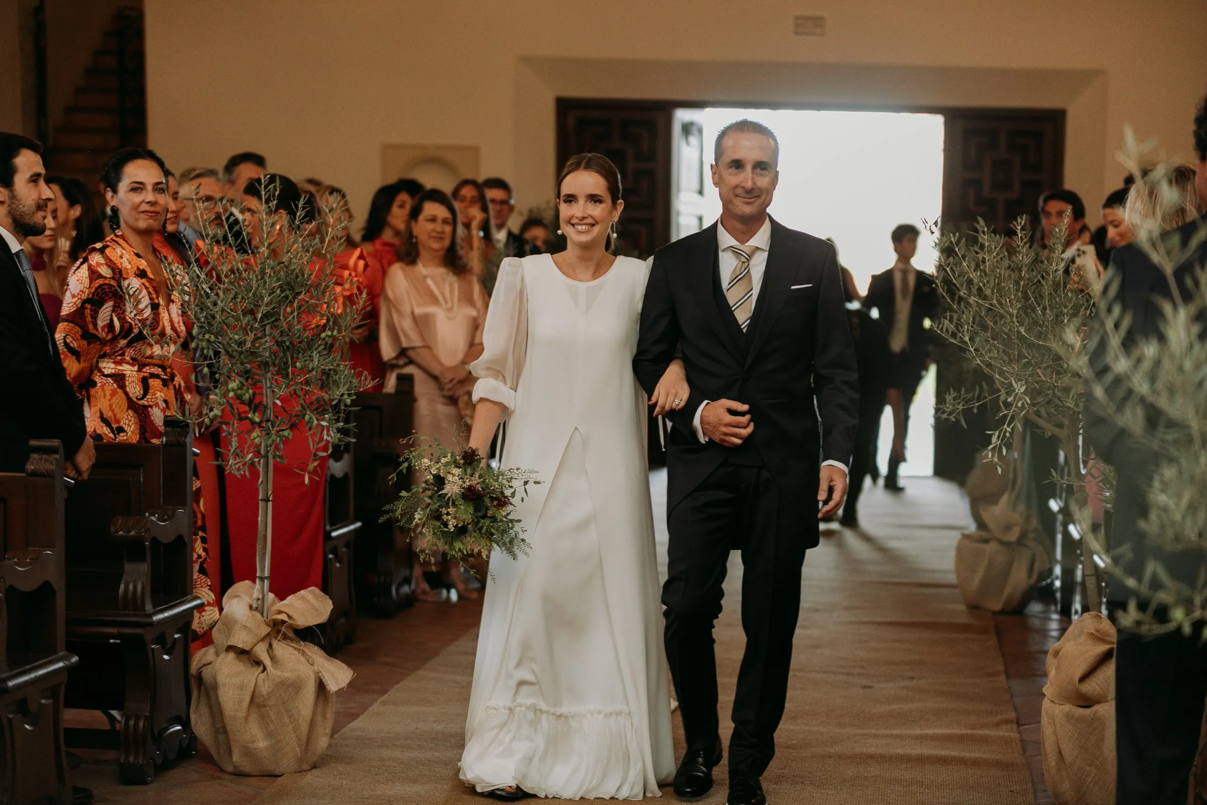 Bride walking down the aisle on her father's arm between olive trees, Nuestra Señora de la Merced