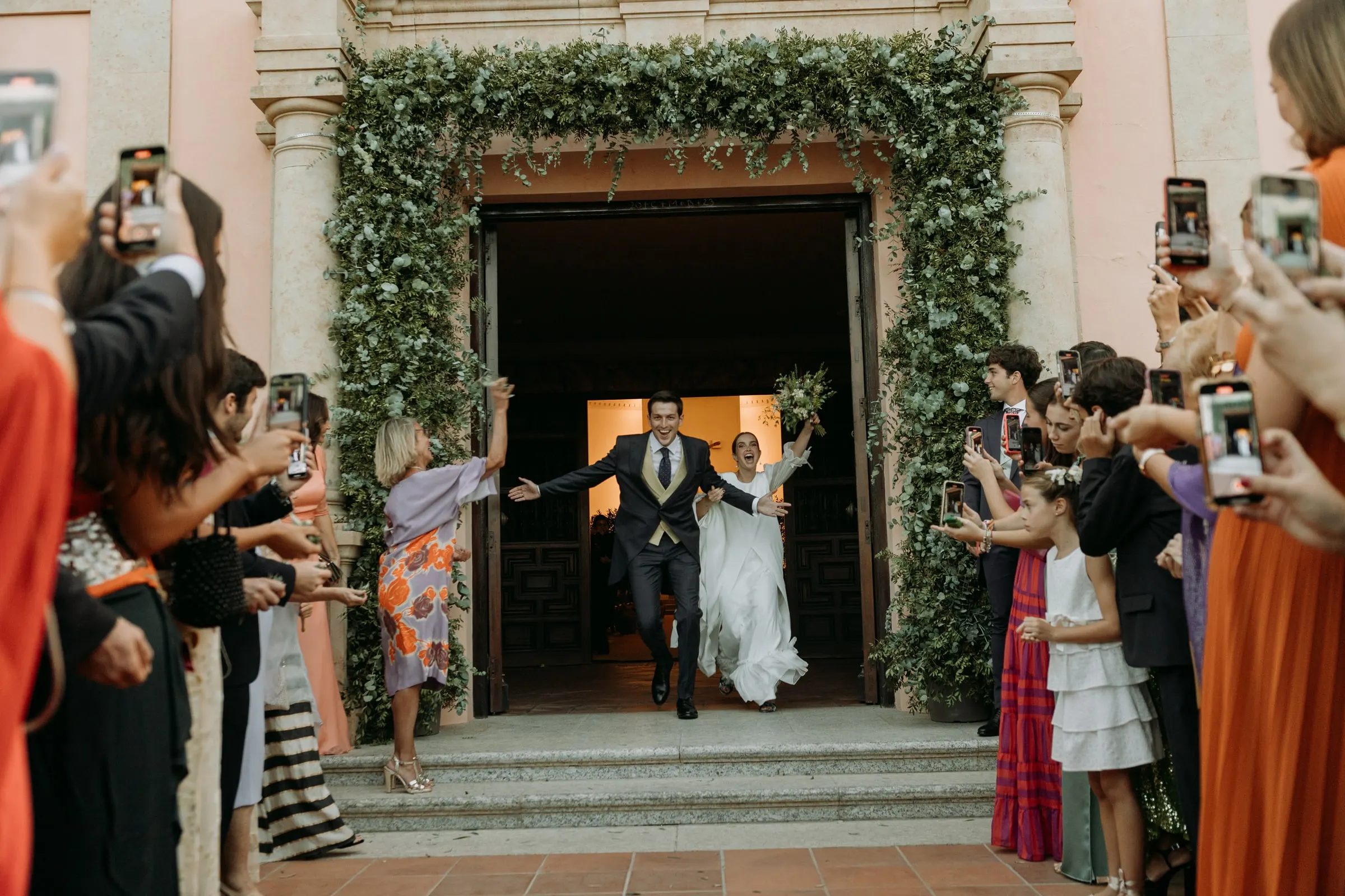 Newlyweds bursting out of the church door framed in greenery as guests film them, Sotogrande