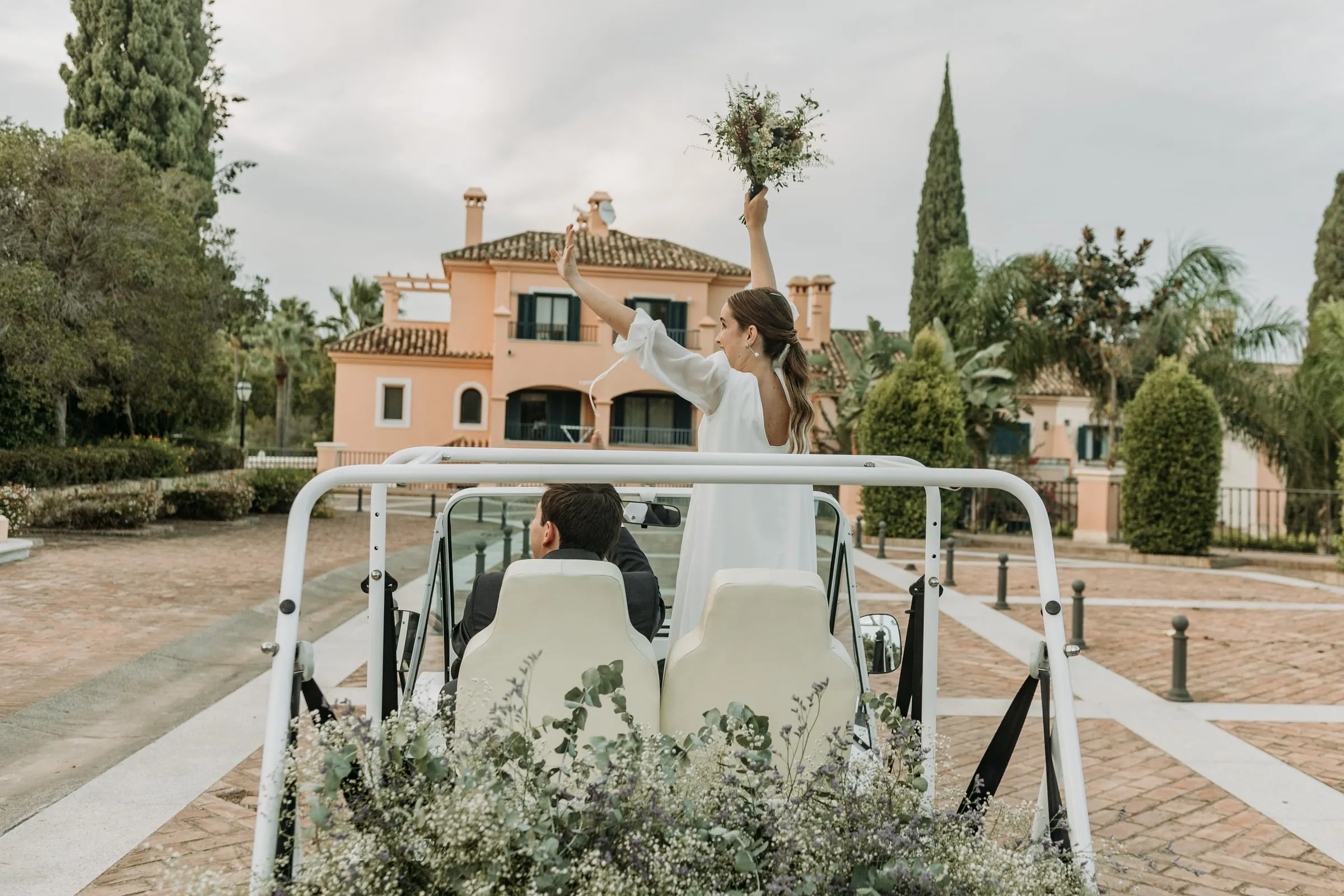 Bride standing in the open car waving her bouquet as they drive off through Sotogrande