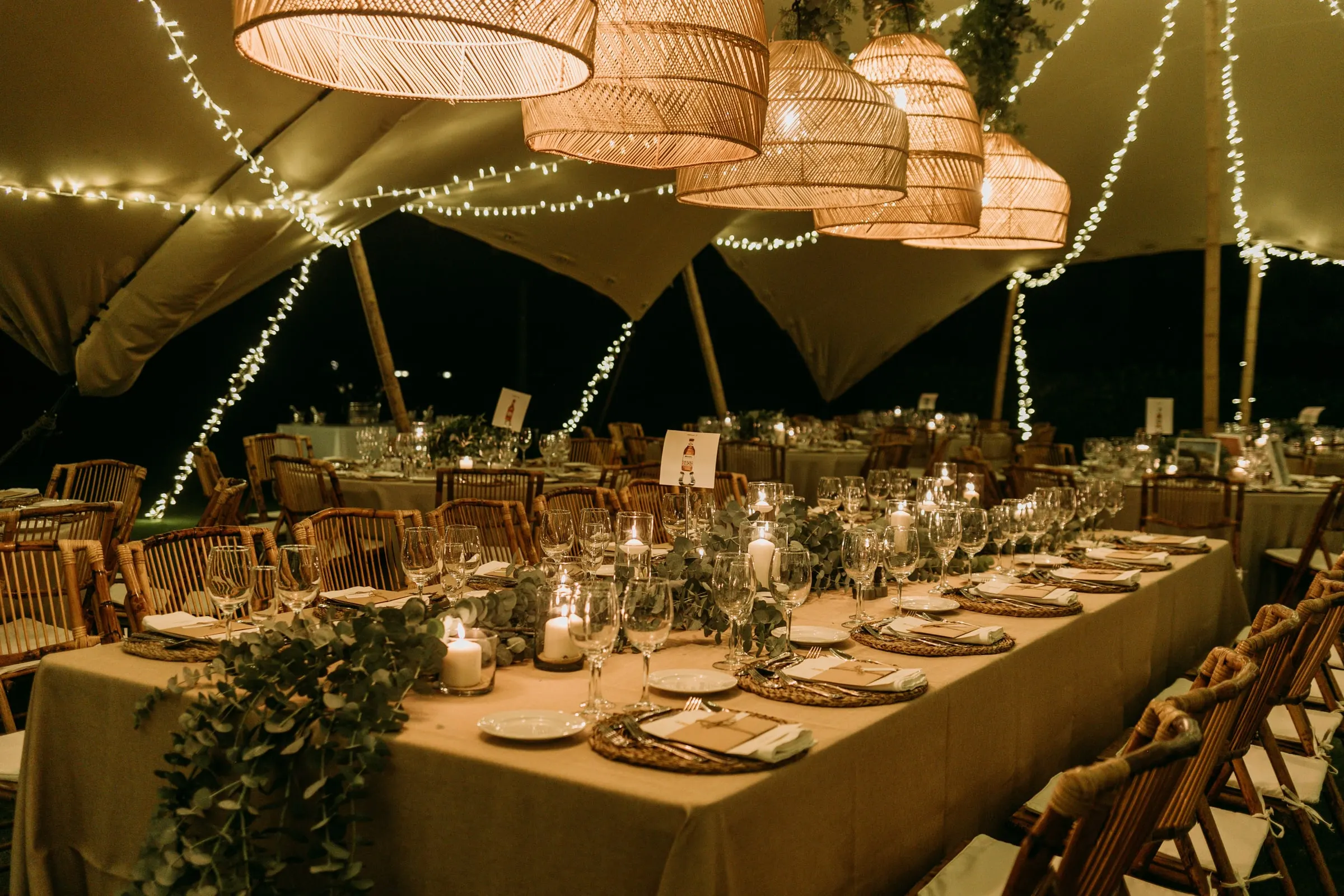 Long banquet tables under a tent with rattan lamps and fairy lights