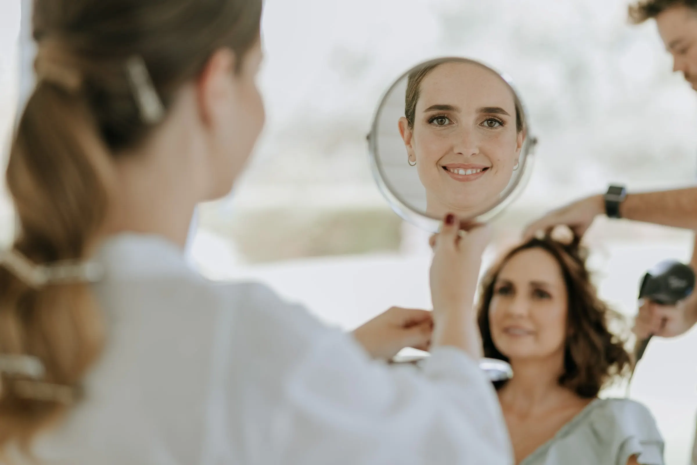 Bride's face reflected in a hand mirror while another woman has her hair done behind her