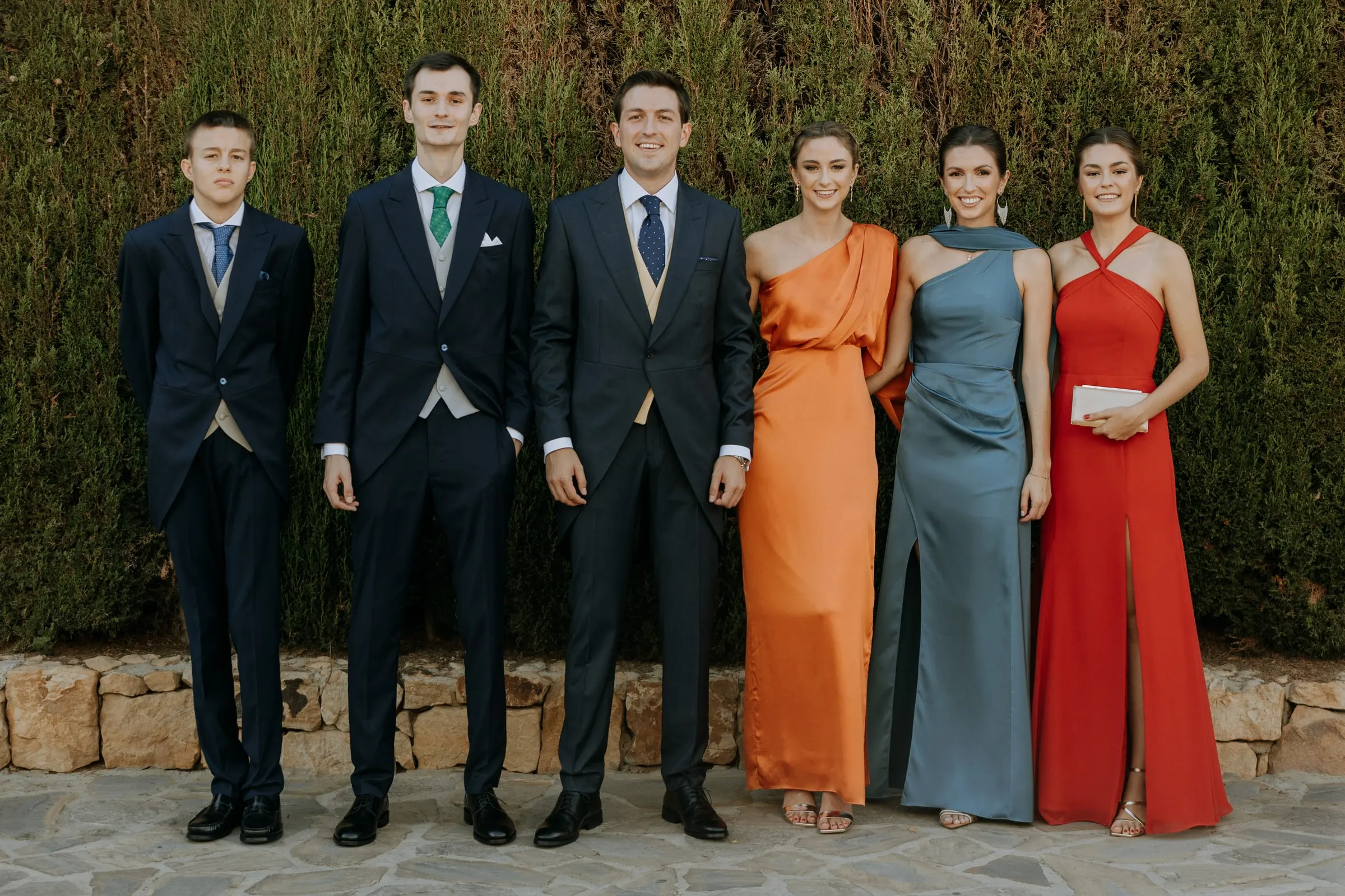 Groomsmen in morning suits and guests in orange, blue and red gowns lined up against the garden hedge in Sotogrande