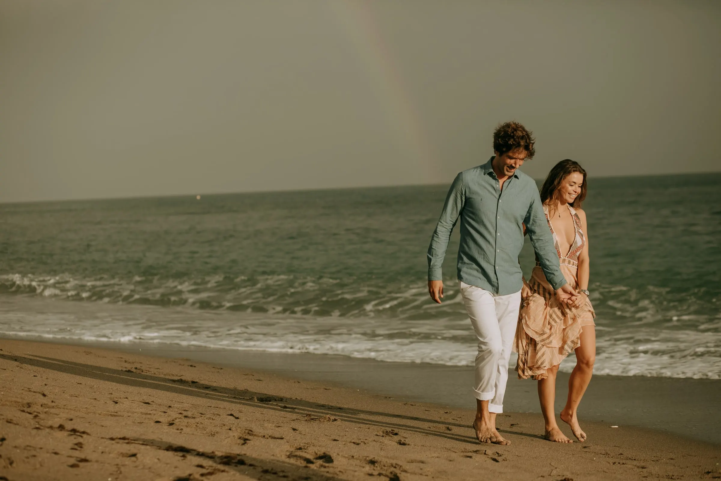 Lauren and Arnaud walking barefoot along the shore under a faint rainbow, pre-wedding session on the beach at Sotogrande