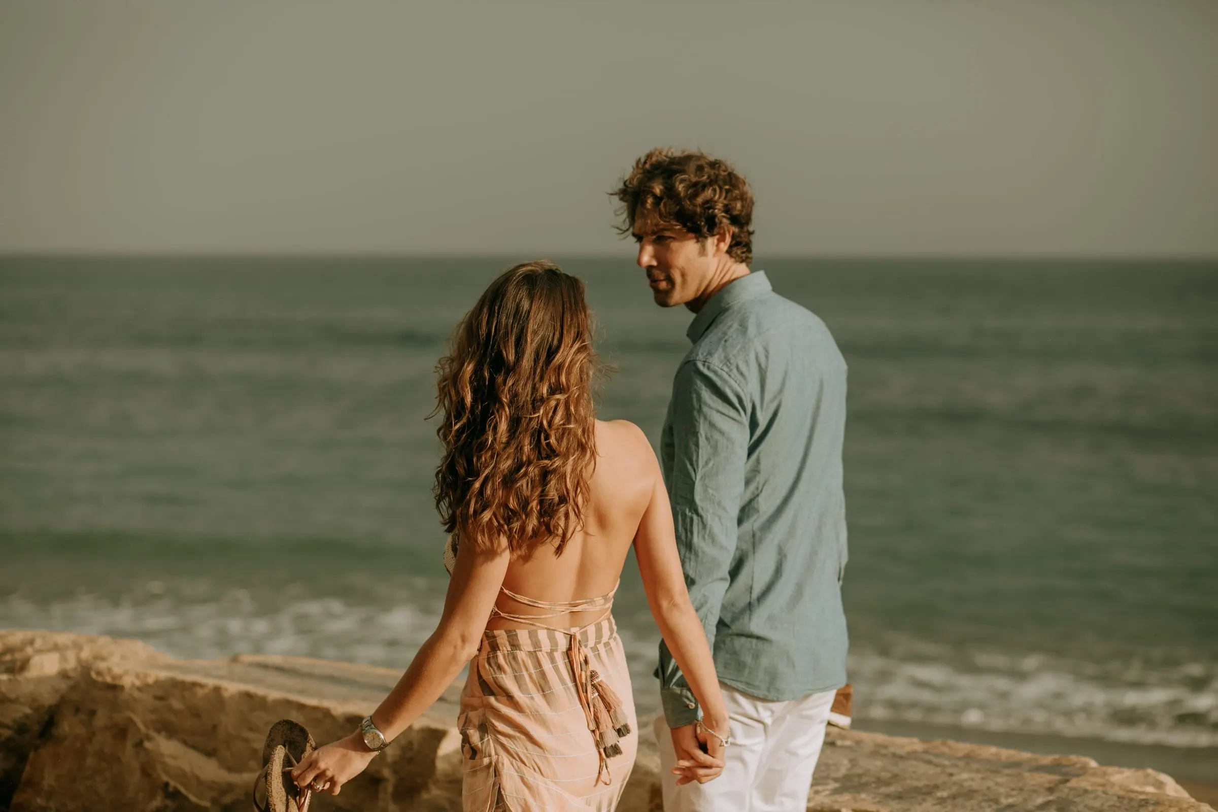Bride-to-be in an open-back pink dress walking to her fiancé on the rocks, pre-wedding on the beach, Sotogrande