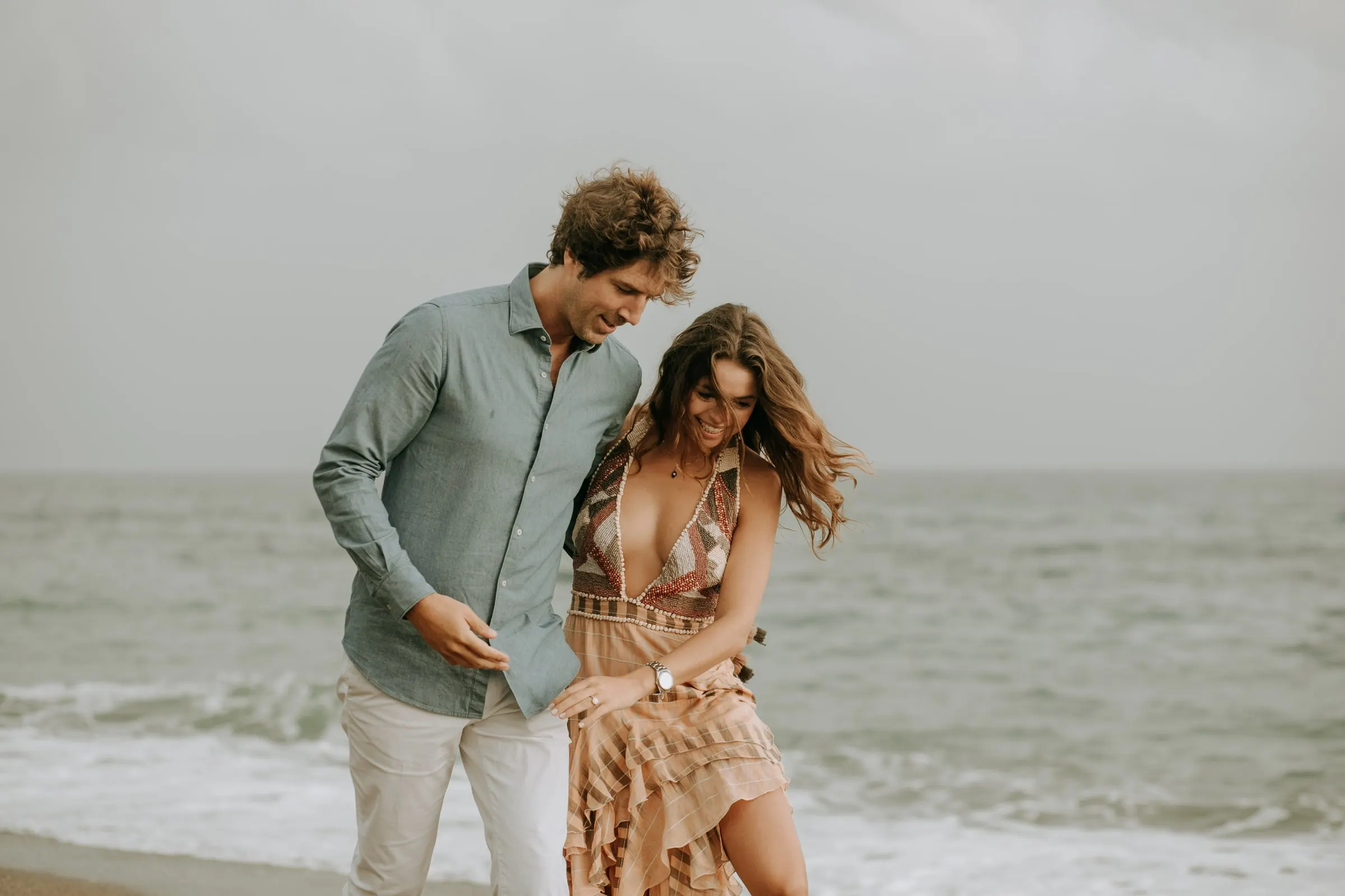 Lauren se ríe apoyada en el hombro de Arnaud, con el pelo al viento, sesión preboda en la playa de Sotogrande