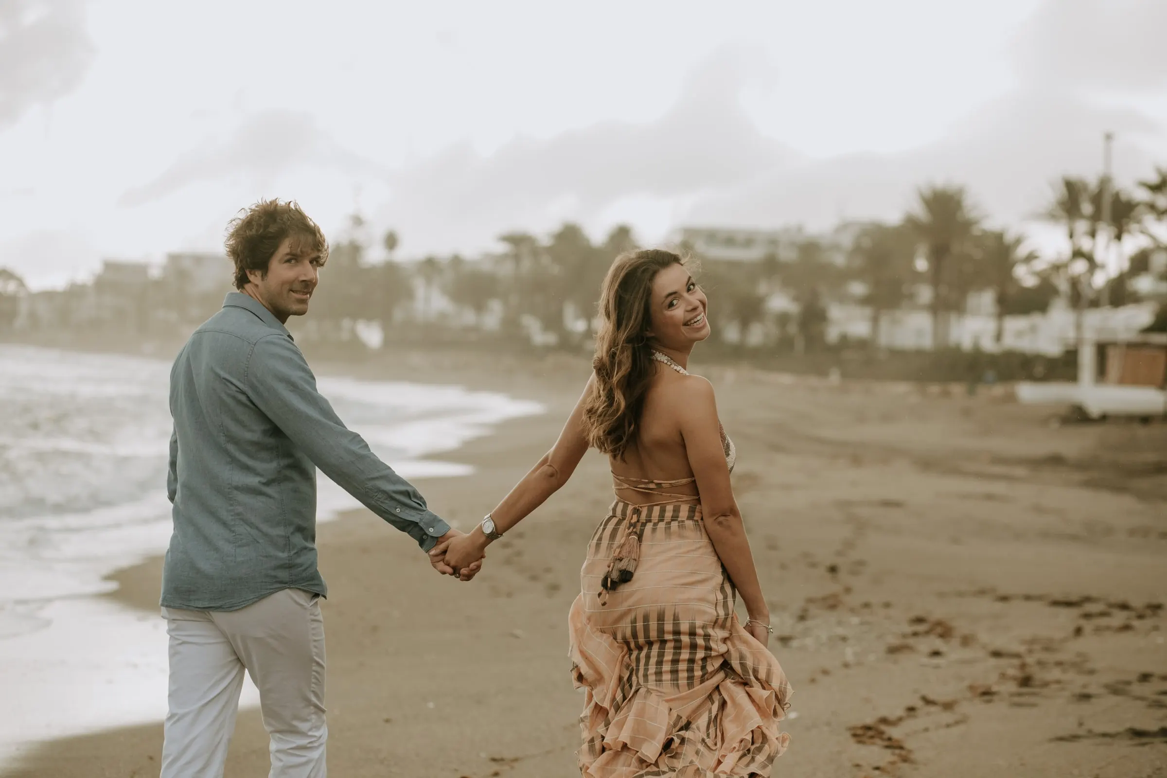Ella le lleva de la mano por la playa y se gira sonriendo, sesión preboda en la playa, Sotogrande
