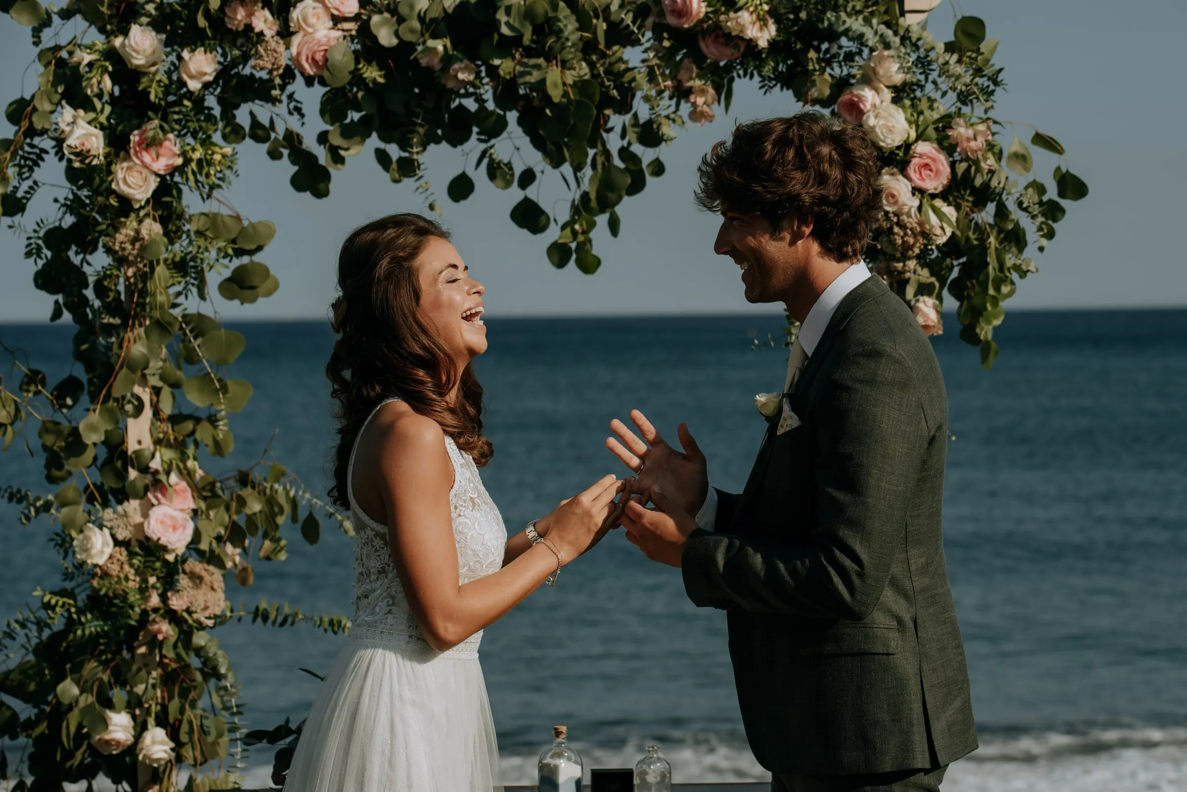 Bride laughing as the groom slides on her ring under the flower arch by the sea, Trocadero, Sotogrande