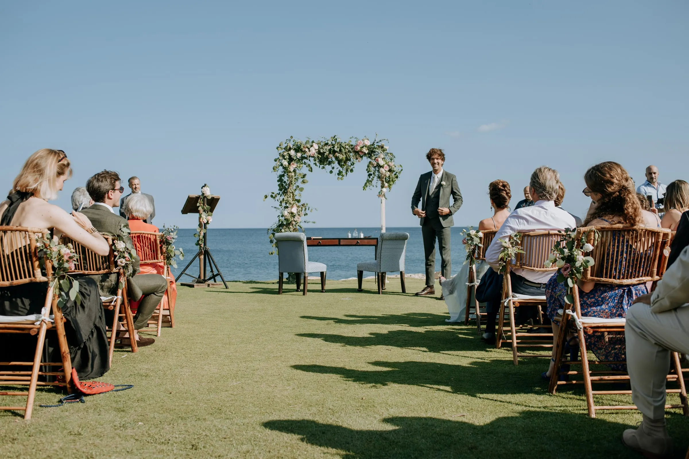 Guests seated on the lawn as the groom waits by the flower arch facing the sea, Trocadero, Sotogrande