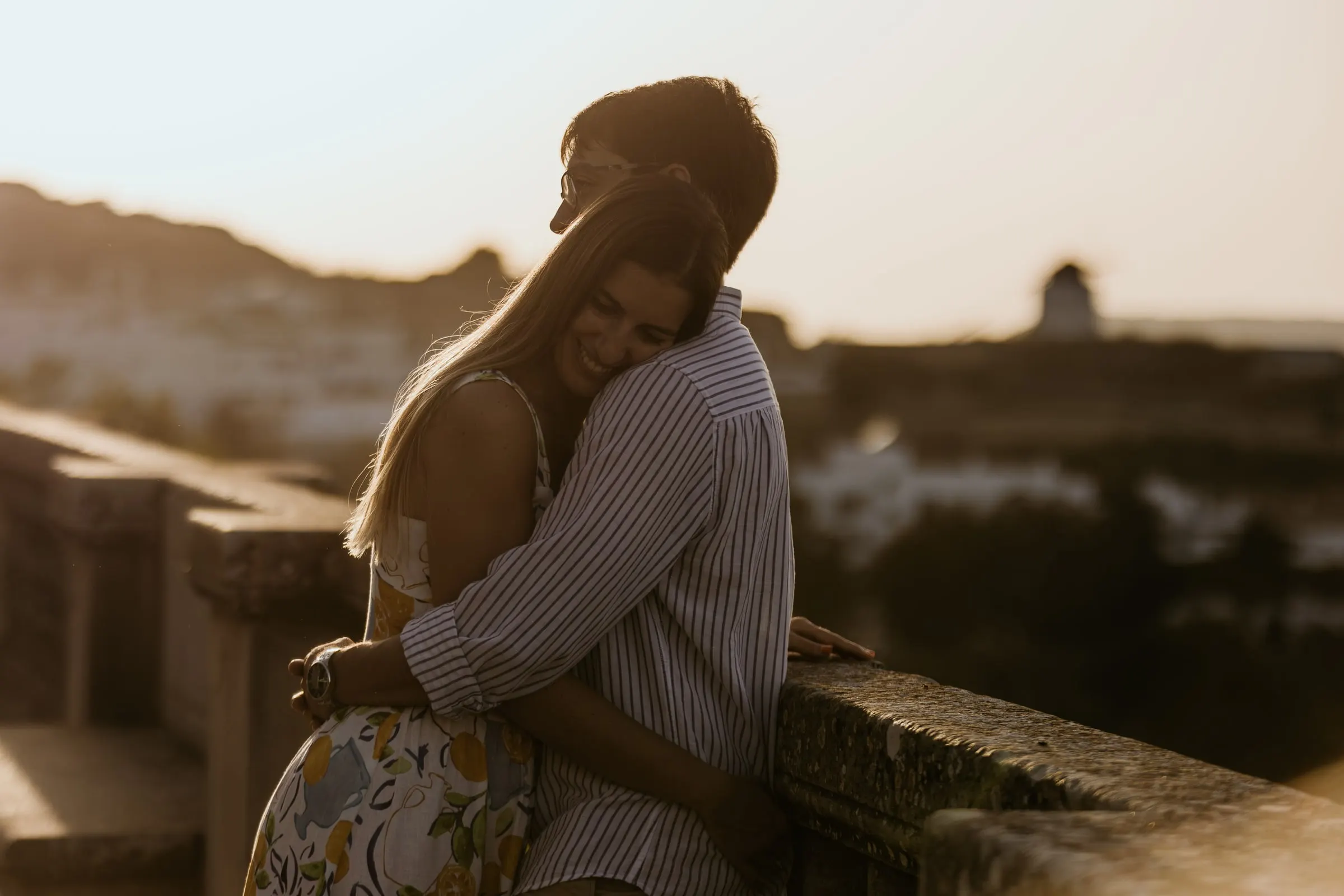 La pareja abrazada en una balaustrada de piedra al atardecer, con un molino en el horizonte de Vejer
