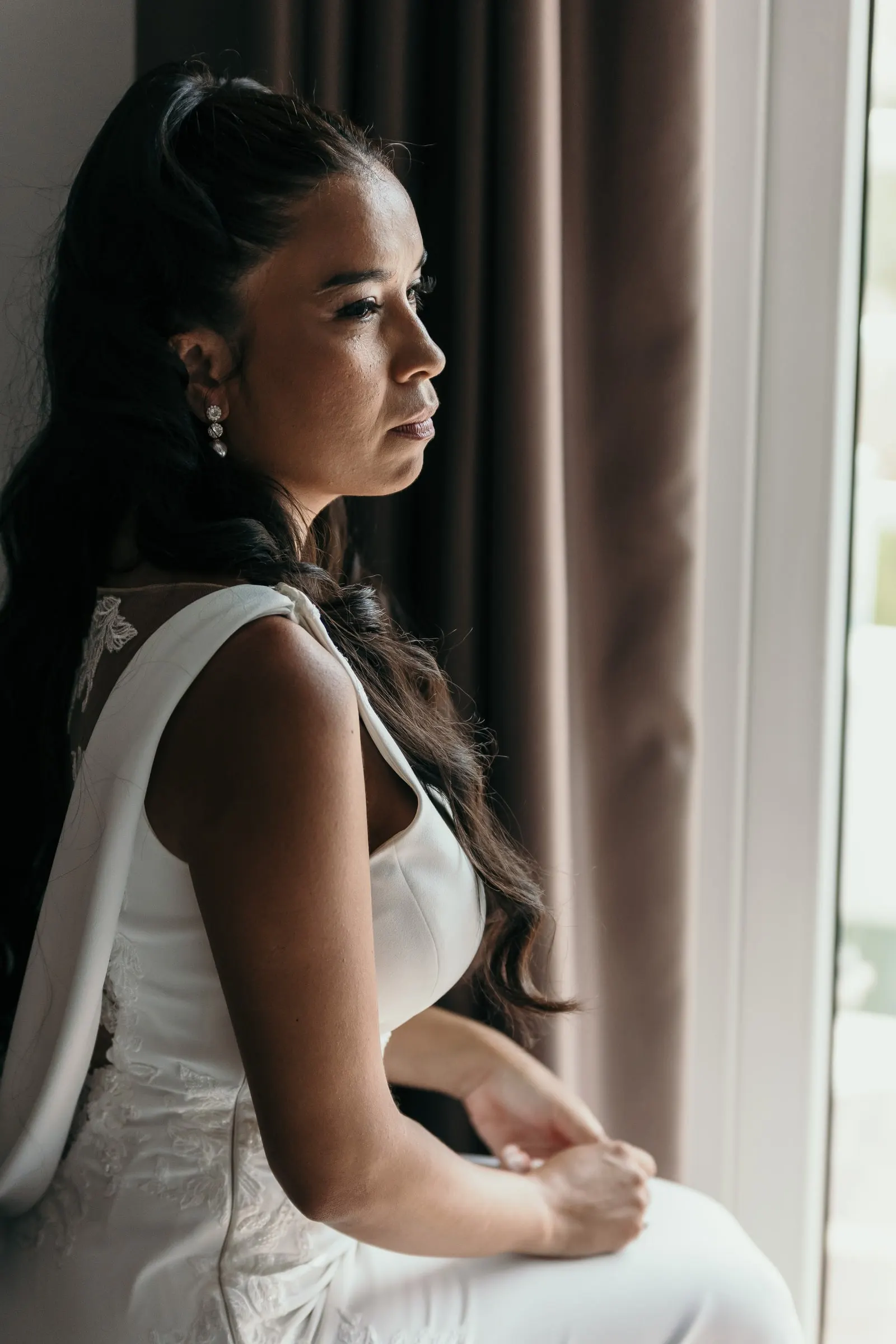 Bride in her wedding dress sitting by the window, looking out in silence