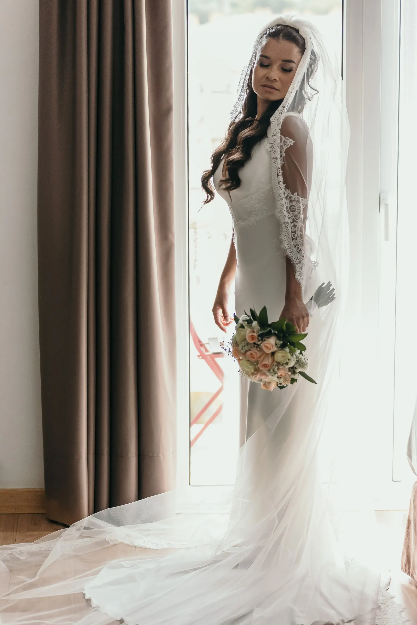 Bride standing in her veil with a bouquet of pink roses by a tall window, Marbella
