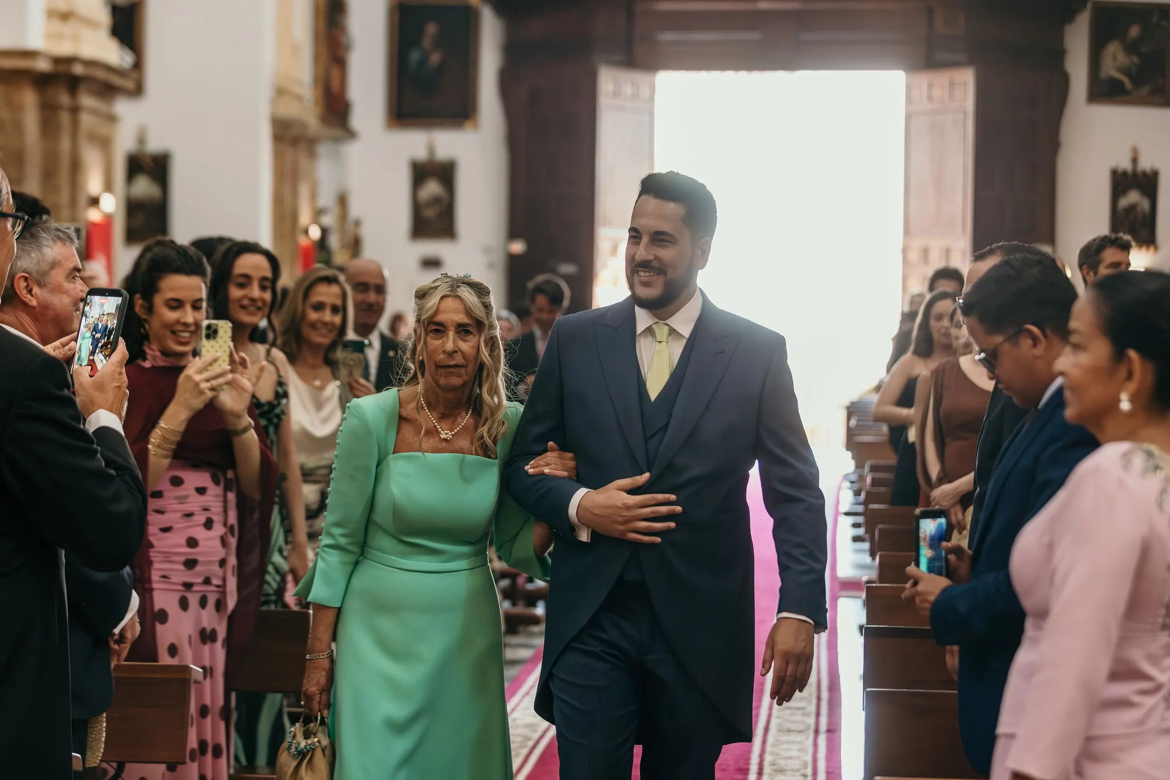 Groom walking down the church aisle arm in arm with his mother in a mint green dress