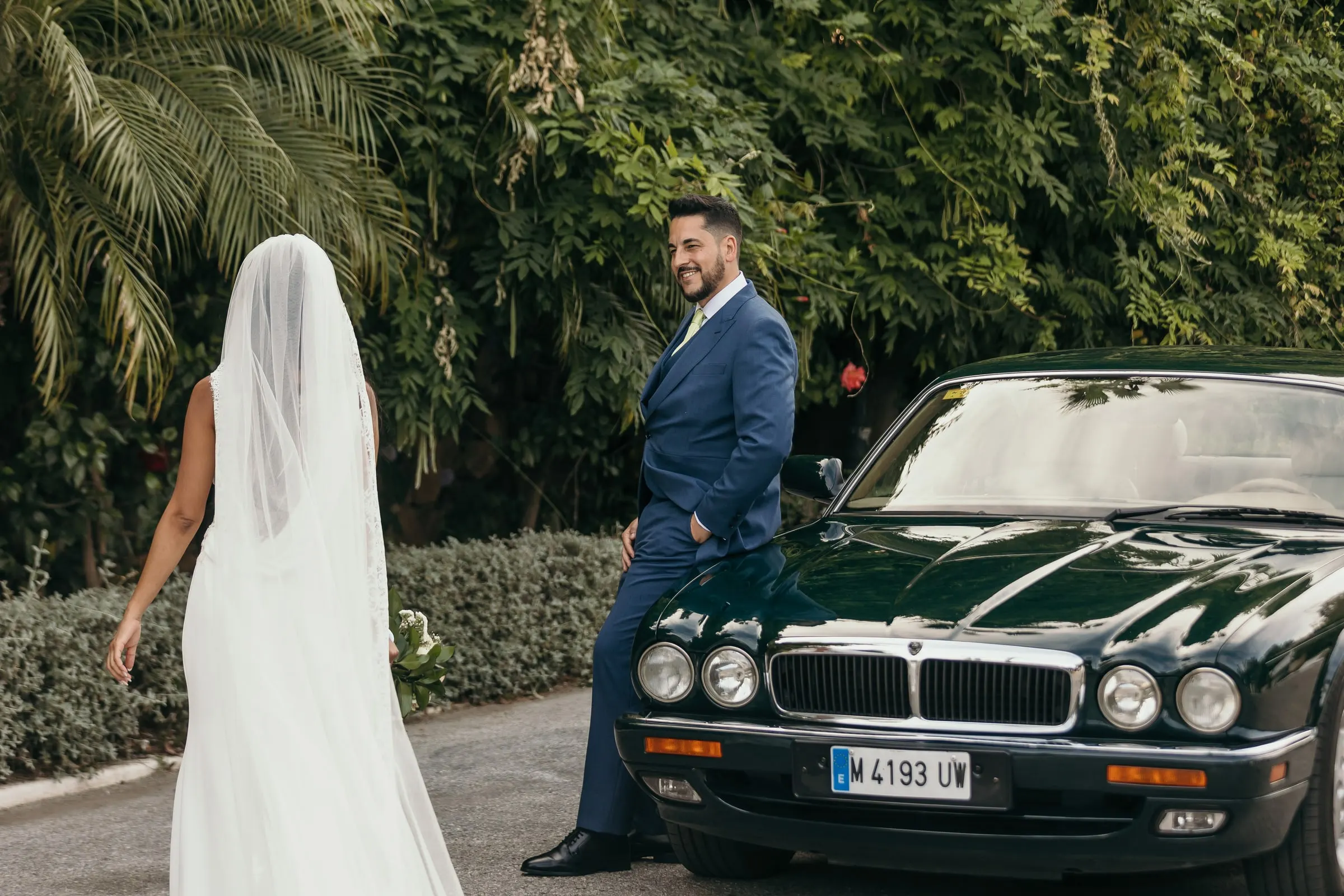 Bride from behind in her long veil walking towards the groom by a classic dark green Jaguar