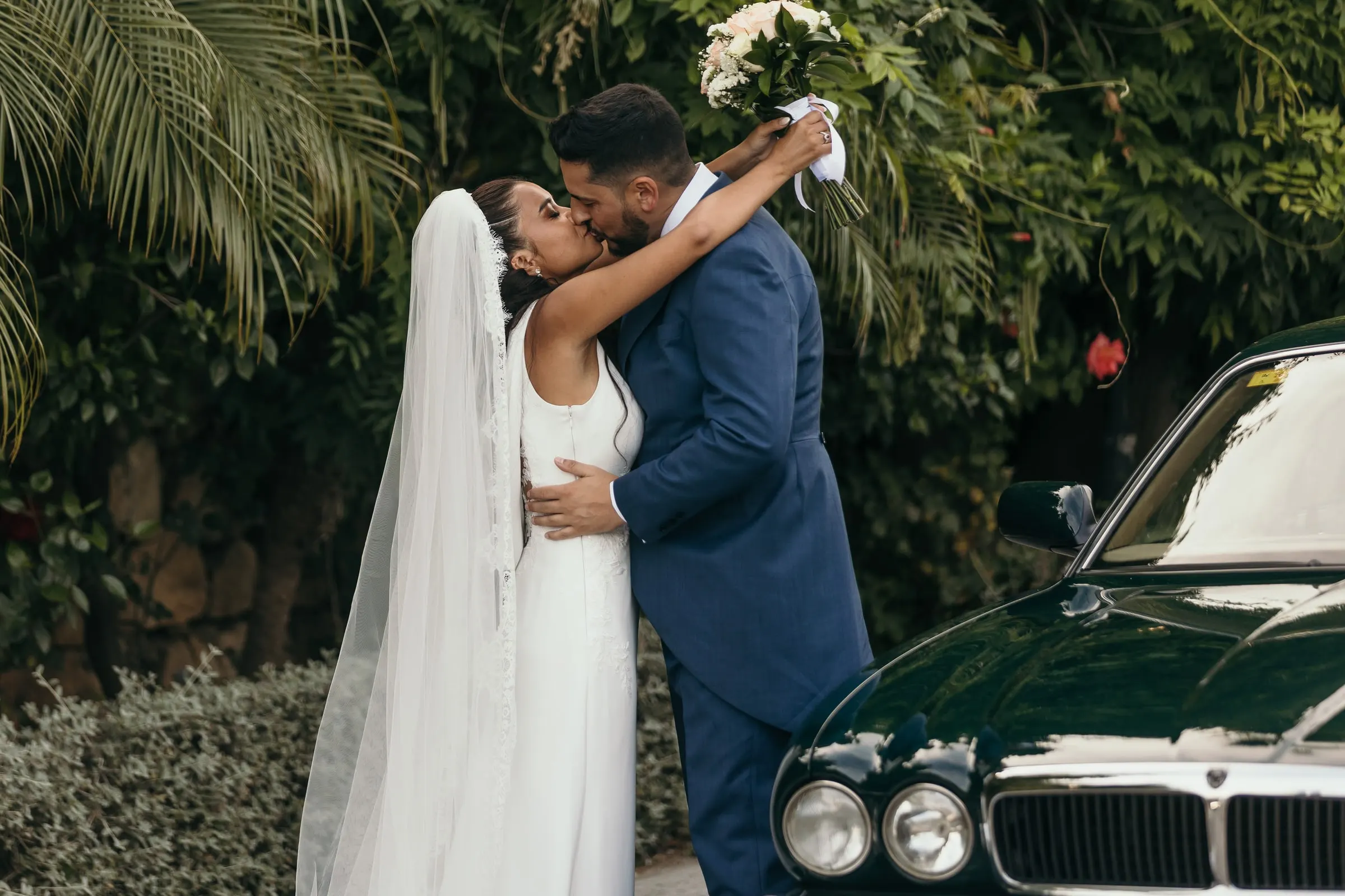 Bride and groom kissing beside the classic wedding car among palms and greenery