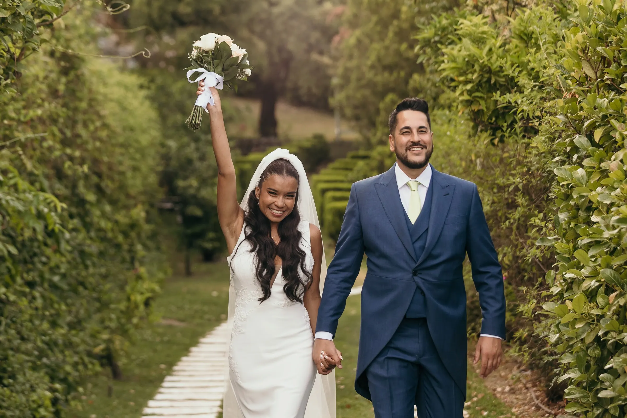 Bride lifting her bouquet in the air as she walks hand in hand with the groom through a garden in Marbella