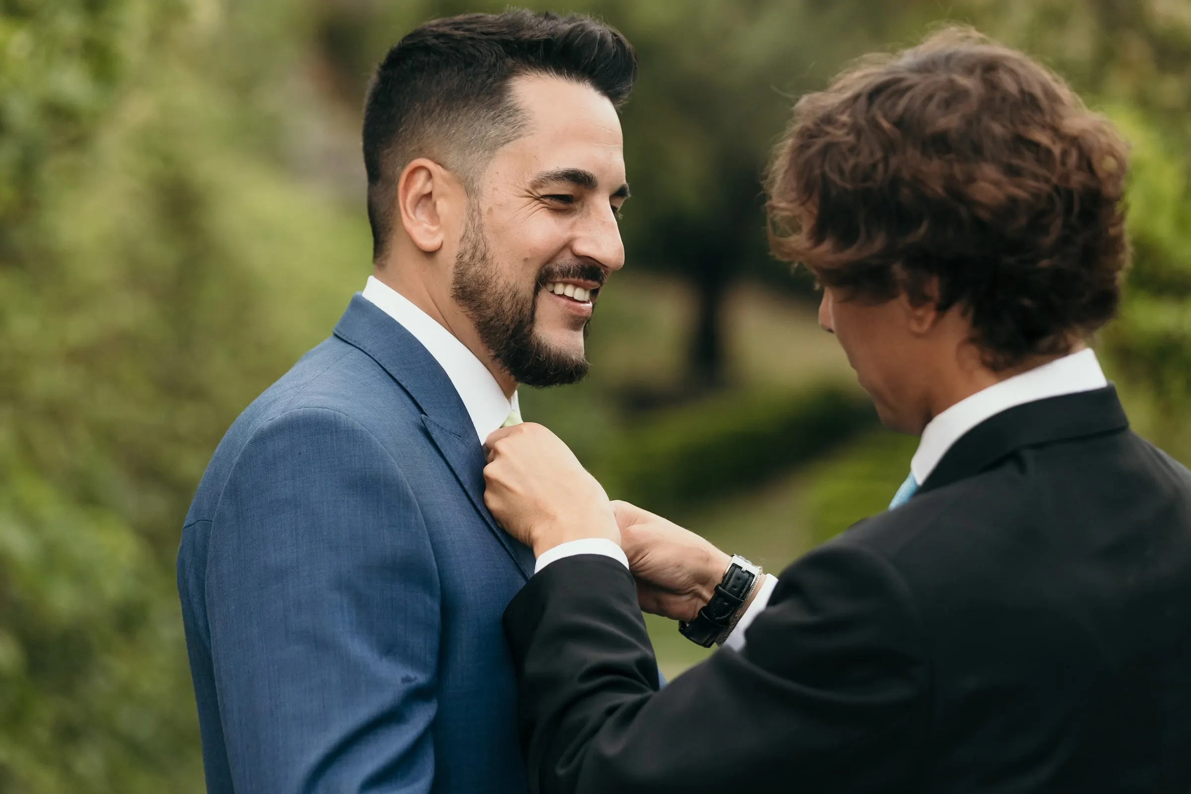 Young guest straightening the groom's tie in the garden, the groom smiling