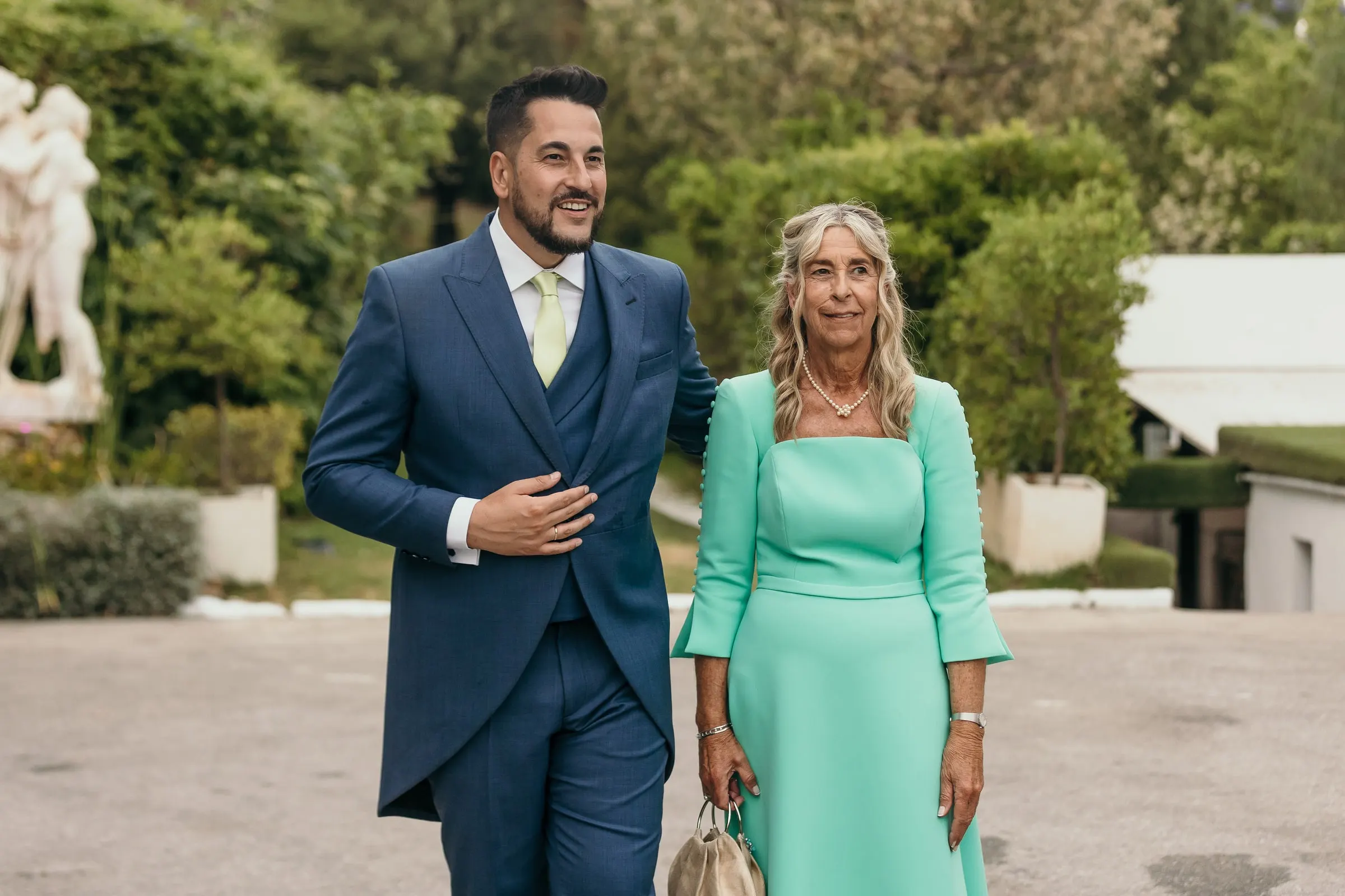 Groom arriving at the reception with his mother past a white marble statue