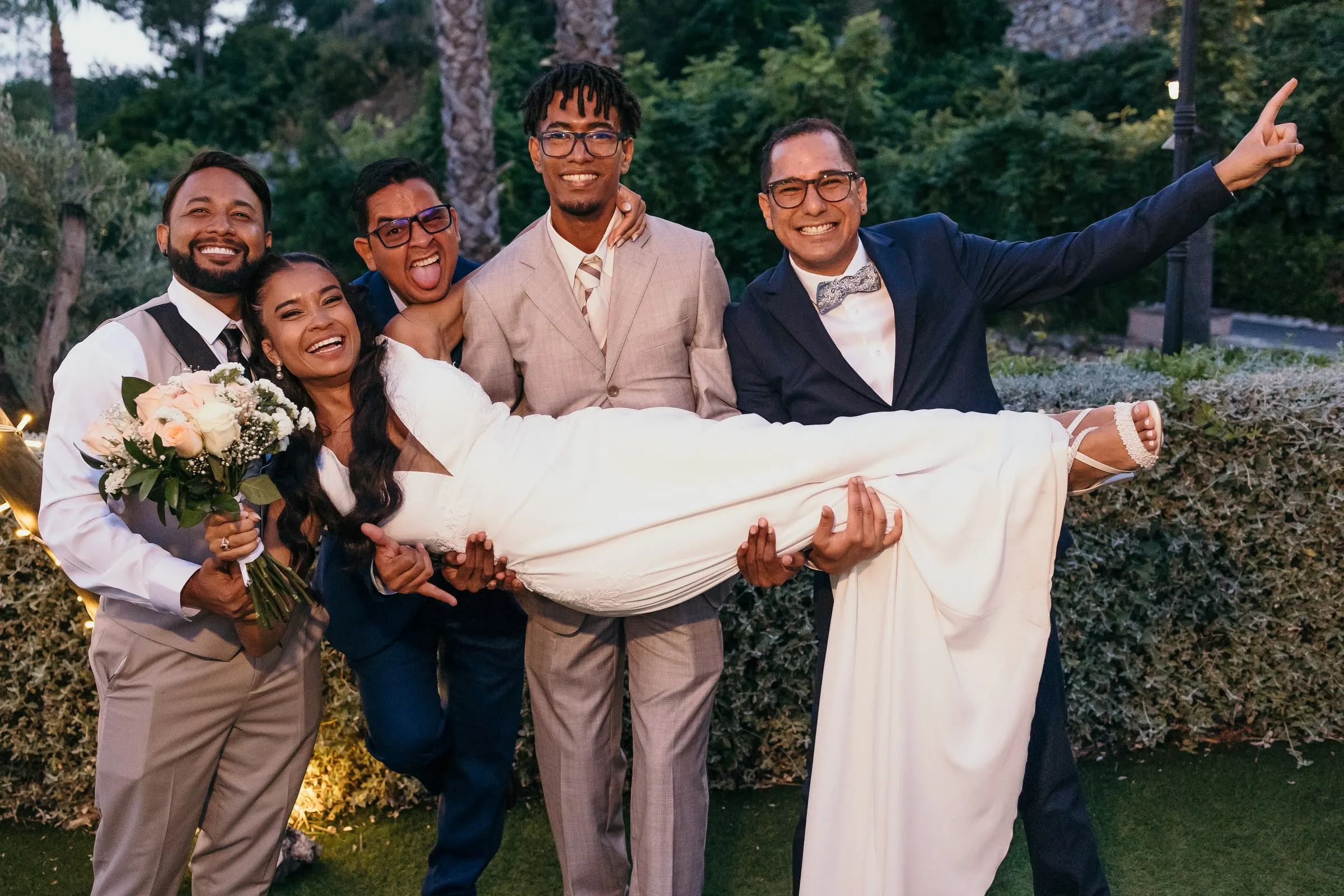 Friends lifting the bride in their arms in the garden at dusk