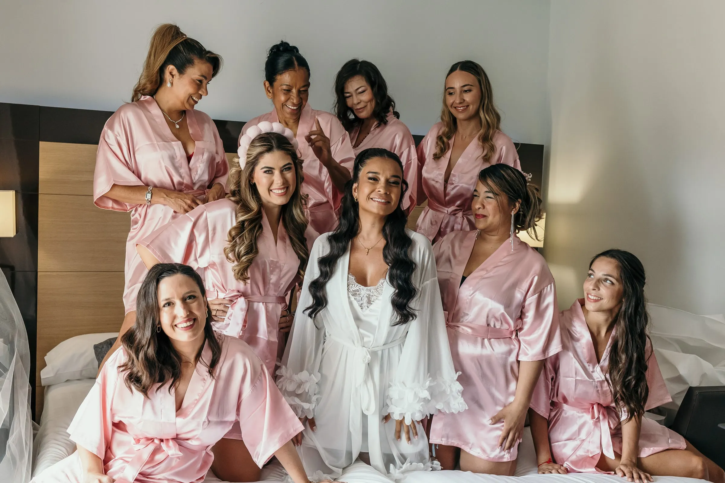 Bride in a white robe sitting on the bed among her bridesmaids in pink satin robes, getting ready in Marbella